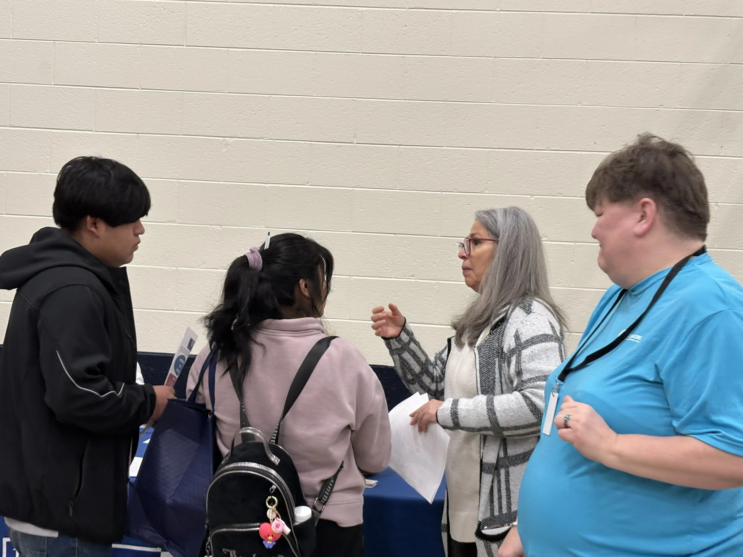 Four people having a conversation indoors, with a beige wall in the background. One woman with gray hair and glasses is speaking, while the others, two men and a woman, listen attentively.