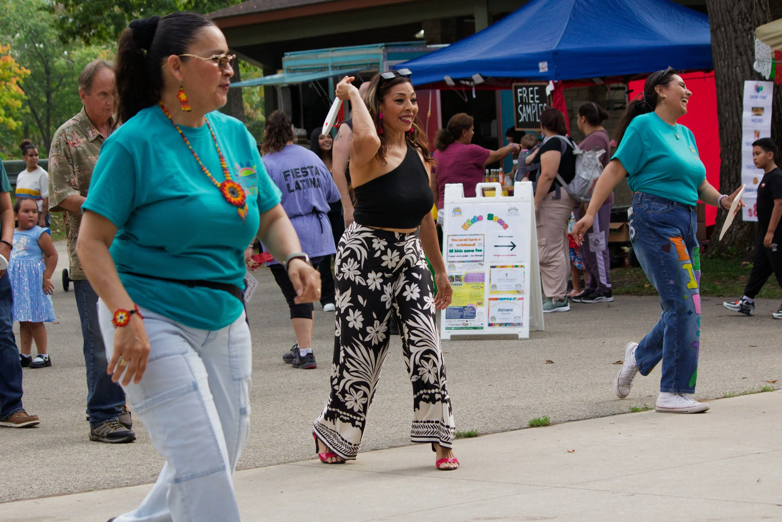 People dancing outdoors at a community event with tents, signs, and children in the background.