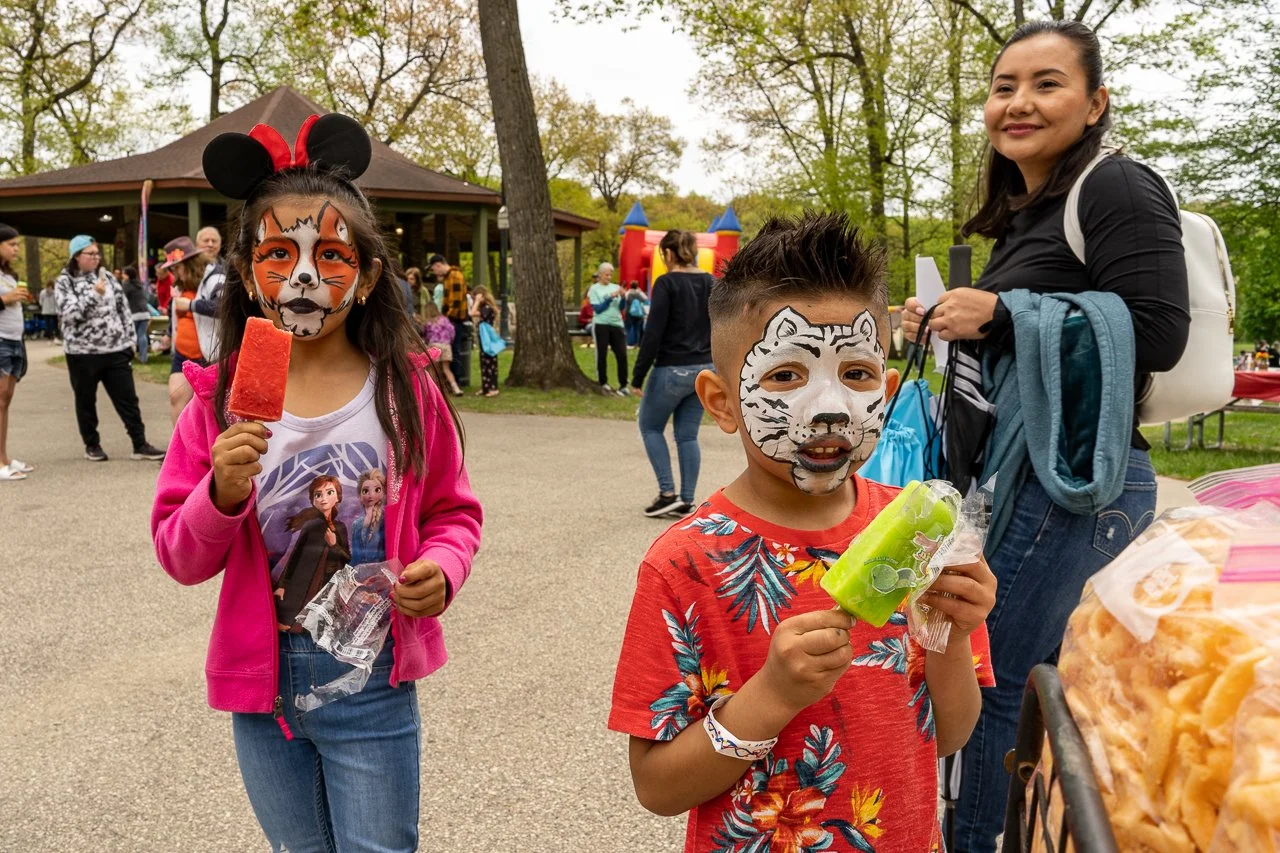 Children with tiger and zebra face paint holding popsicles at an outdoor fair or festival with people, trees, benches, and a bounce house in the background.