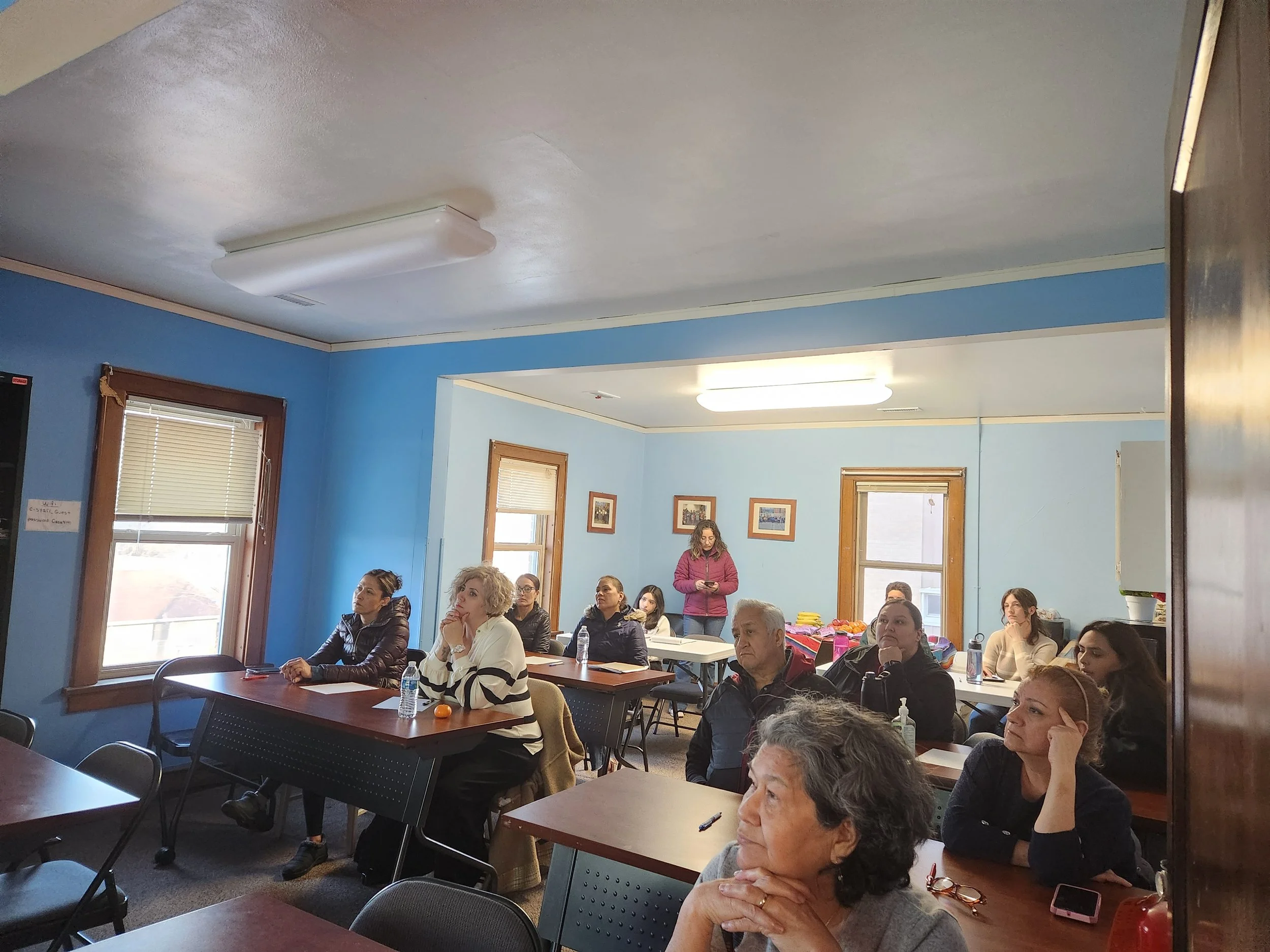 A group of adults attending a meeting or class in a room with blue walls and wooden trim, sitting at tables with notebooks and water bottles, some looking towards the front and one woman standing and looking at her phone.