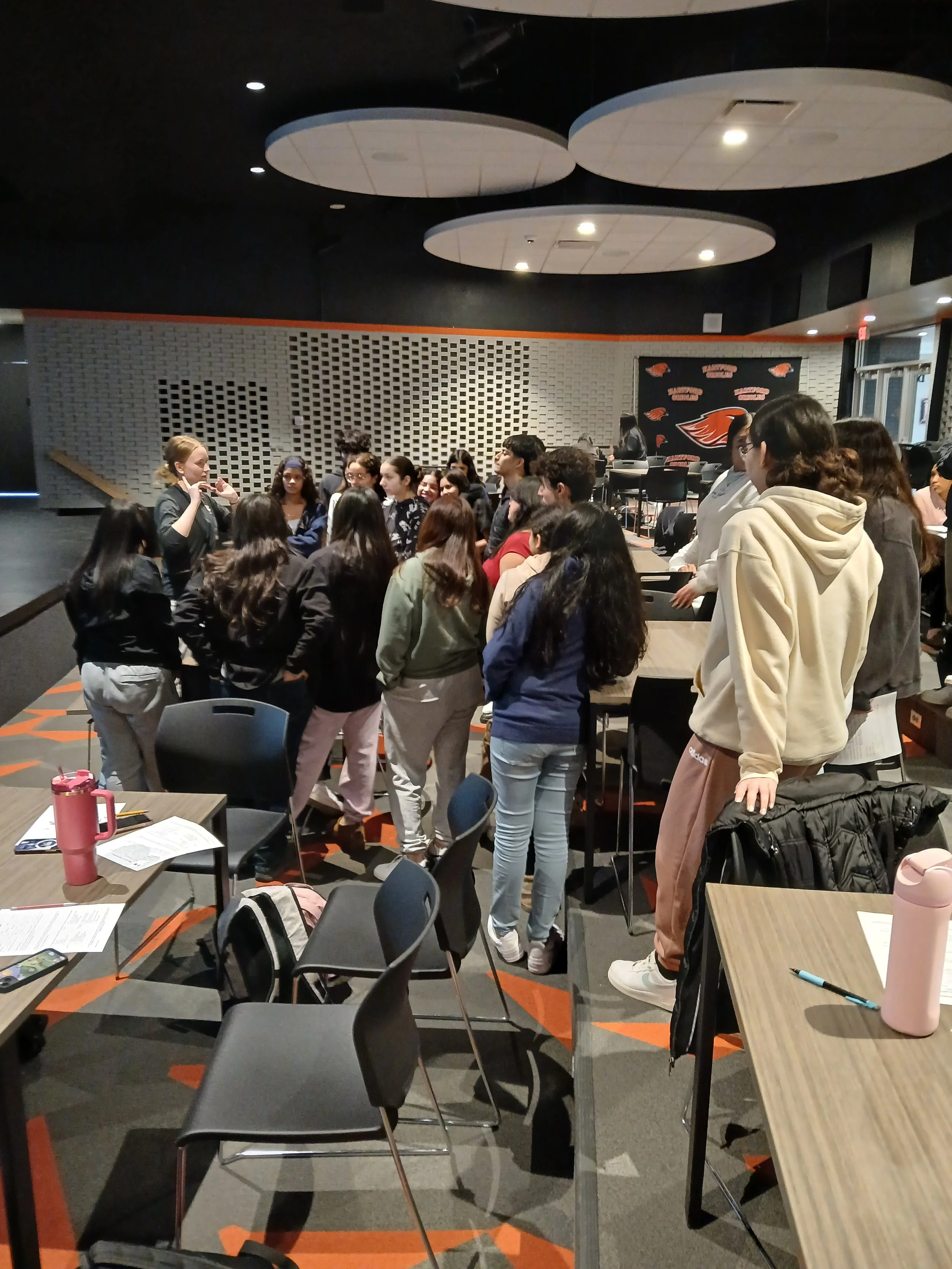 Group of people gathered around a speaker in a conference room with papers, water bottles, and chairs visible.
