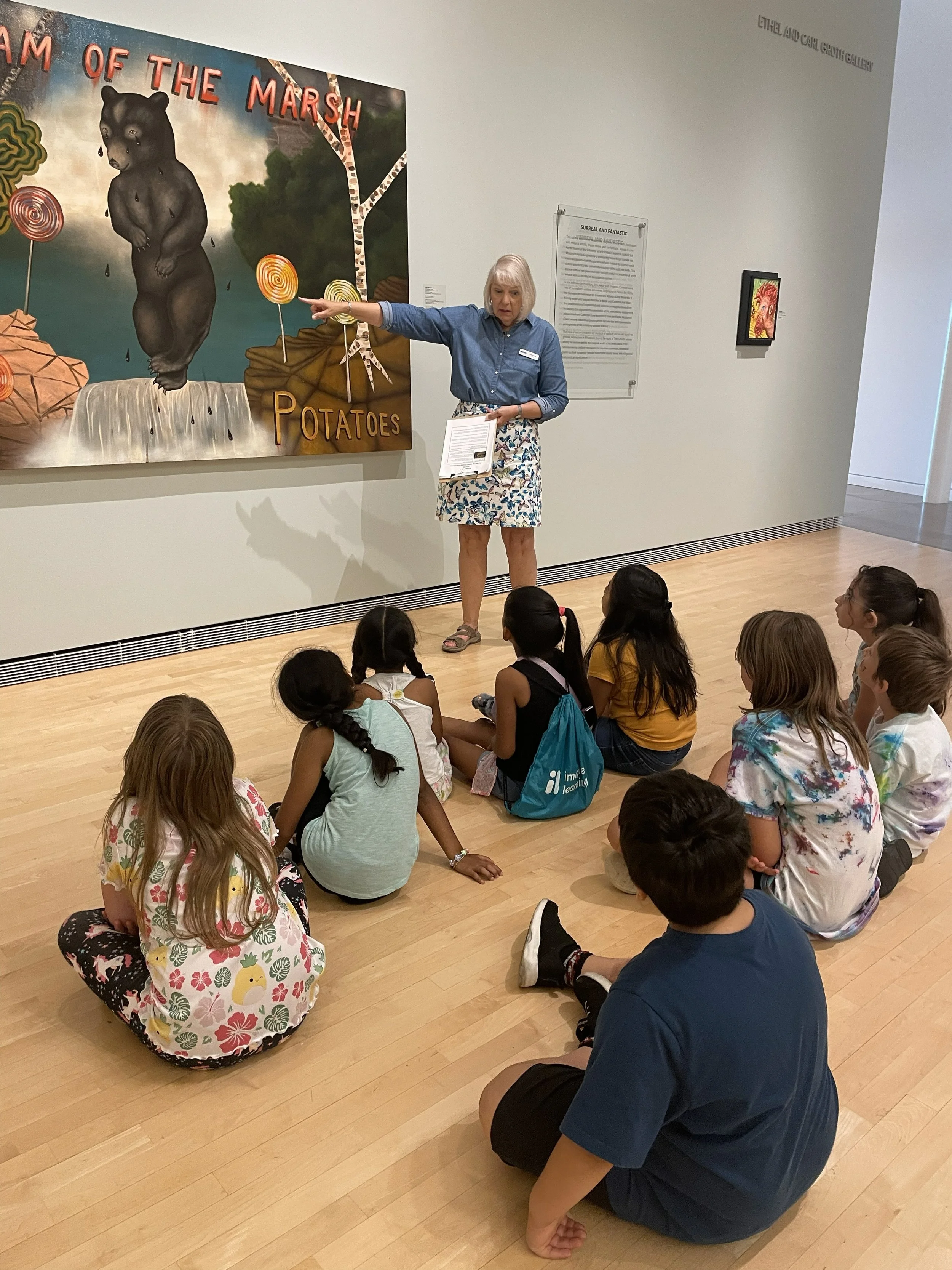 A museum guide is giving a demonstration about a painting titled 'Dream of the Marsh' which features a bear, potatoes, and lollipops. A group of children is sitting on the floor listening.