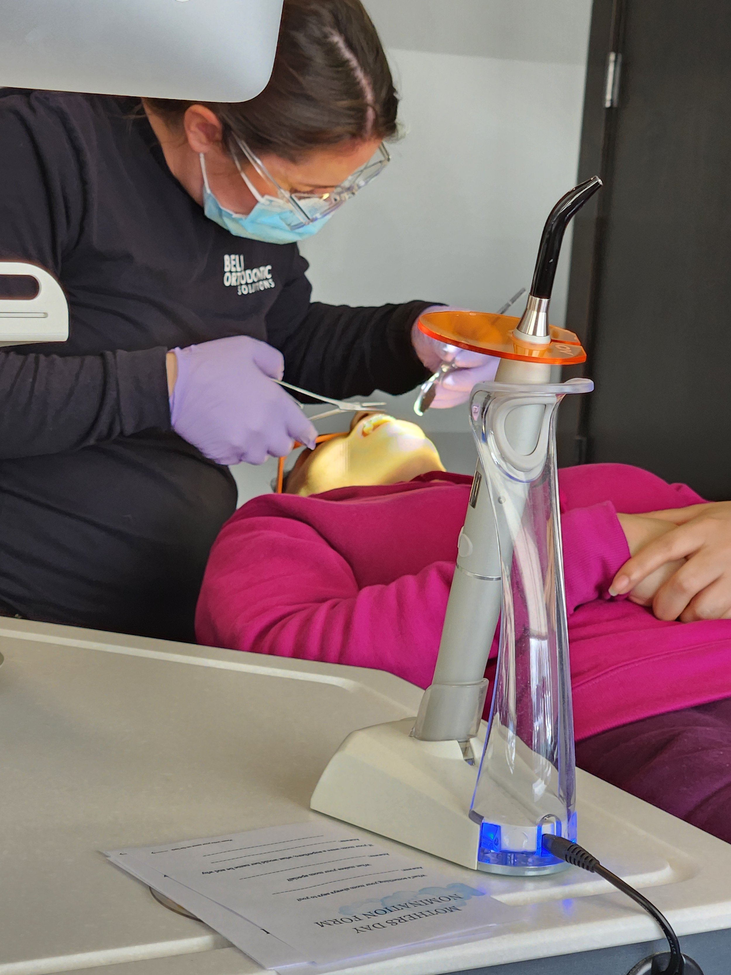 A woman in a pink top is lying on a dental chair receiving a dental procedure from a dental professional. The dental professional is wearing protective gear, including gloves, a mask, and glasses, and is working on the woman's mouth with dental tools. A dental instrument and a water suction device are visible, with the latter connected to a power source.