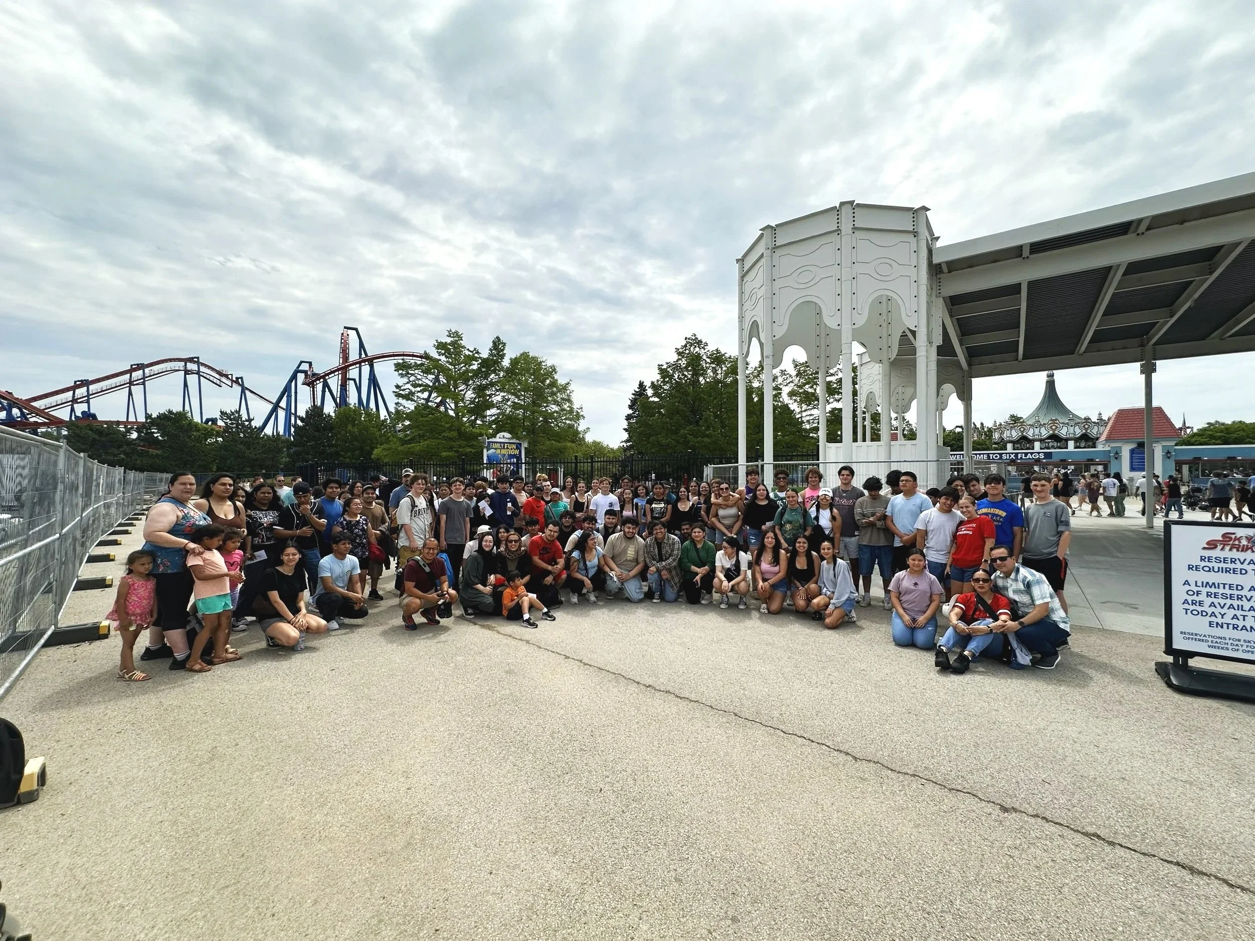 Group of people gathered at an amusement park, with roller coaster tracks in the background and a carousel nearby.