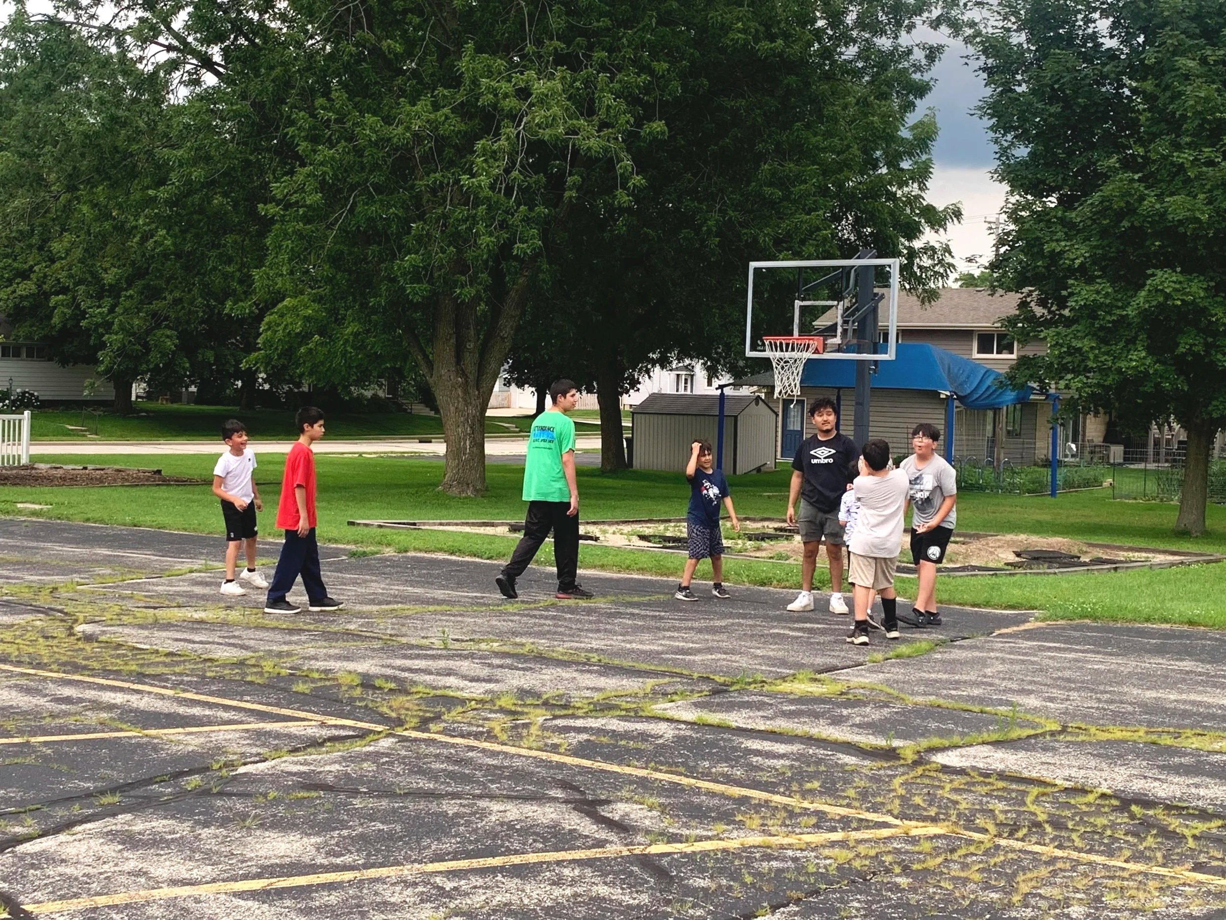 A group of six kids playing basketball on an outdoor court surrounded by trees, with a basketball hoop, residential houses, and cloudy sky in the background.