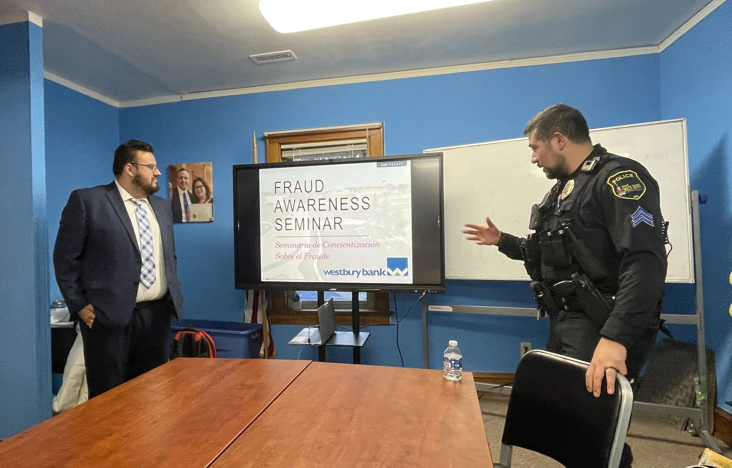 A man in a suit and a police officer are having a discussion in a room with blue walls. A digital screen displays a presentation titled 'Fraud Awareness Seminar' in English and Spanish, with Westbury Bank's logo. There are framed pictures on the wall and a water bottle on the table.