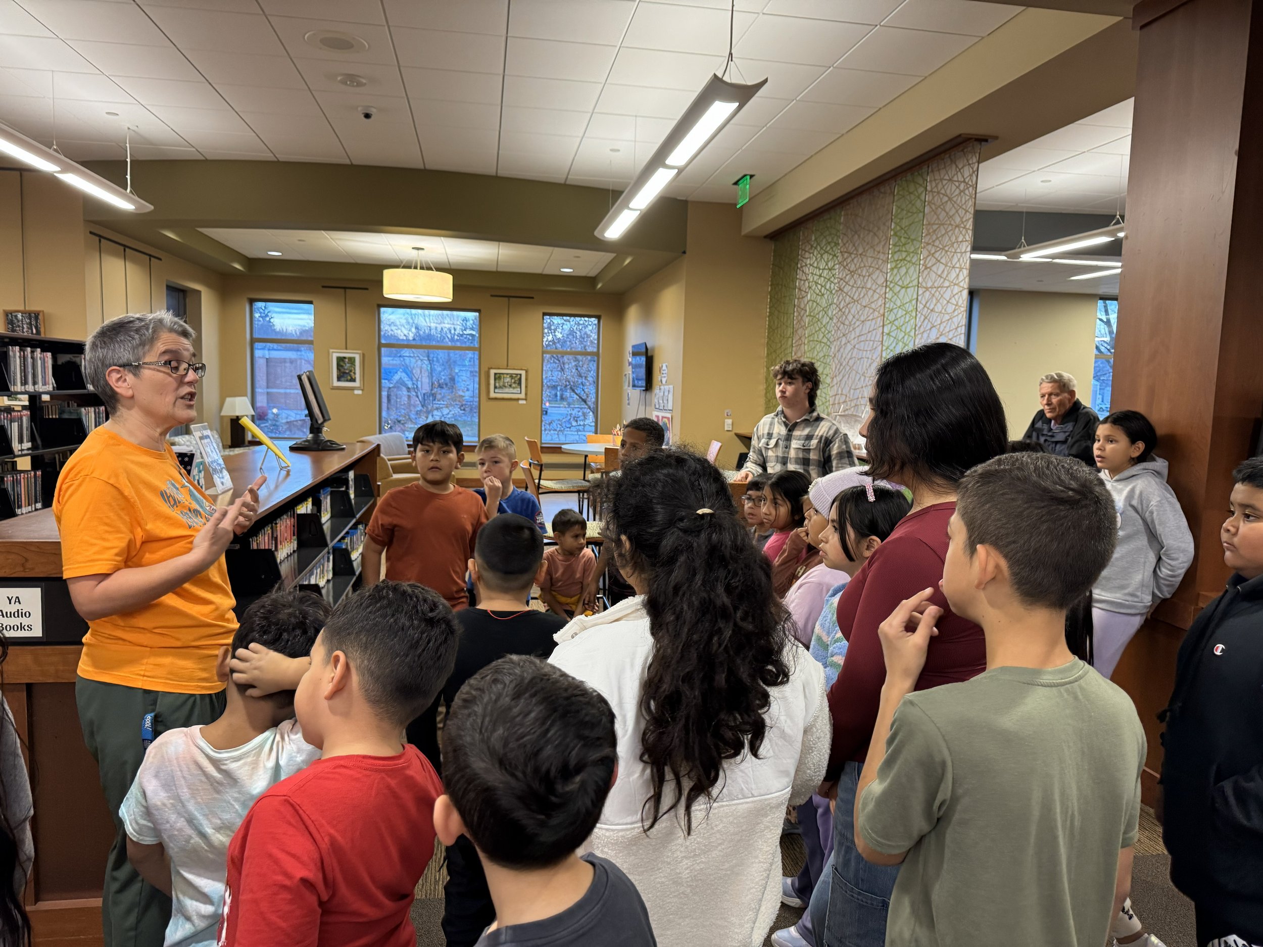 A woman in an orange shirt is speaking to a large group of children and adults in a library, with bookshelves and windows in the background.