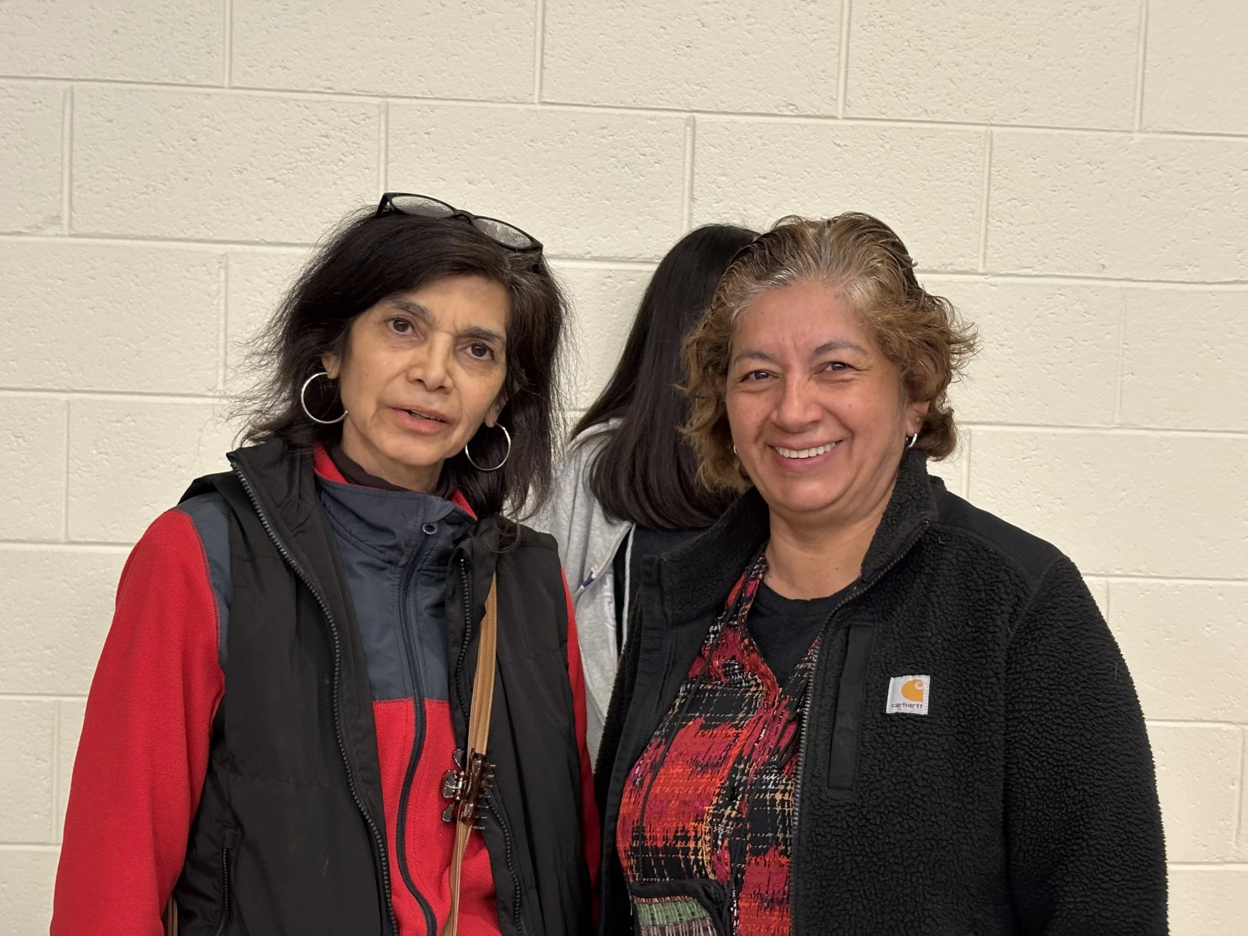 Two women standing in front of a beige brick wall, one with black hair, glasses on her head, wearing a red and black jacket, and the other with curly gray hair, smiling, wearing a black Carhartt fleece jacket and a colorful top.