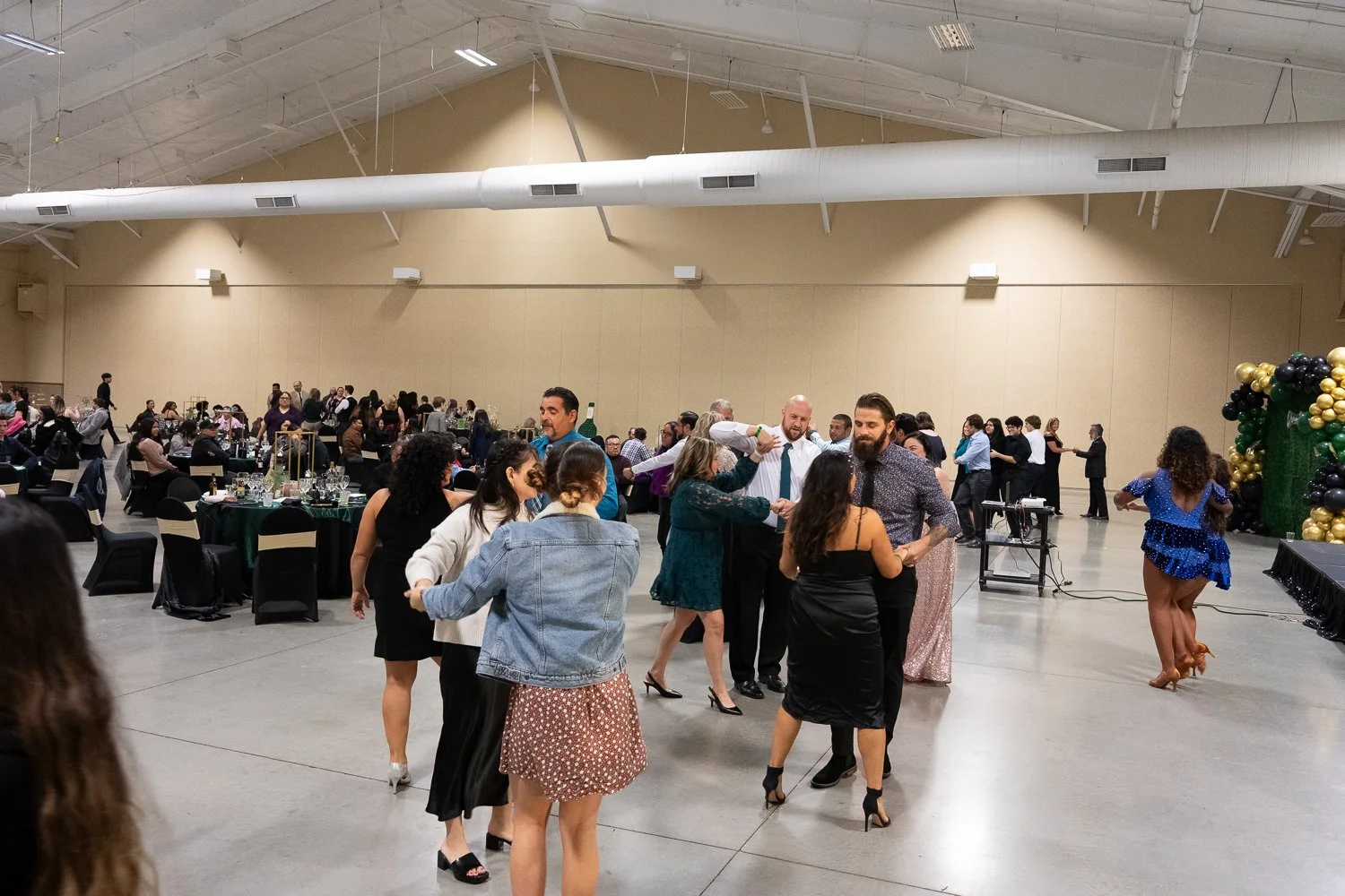 People dancing and socializing at a formal event in a large indoor hall decorated with black, gold, and green balloons.