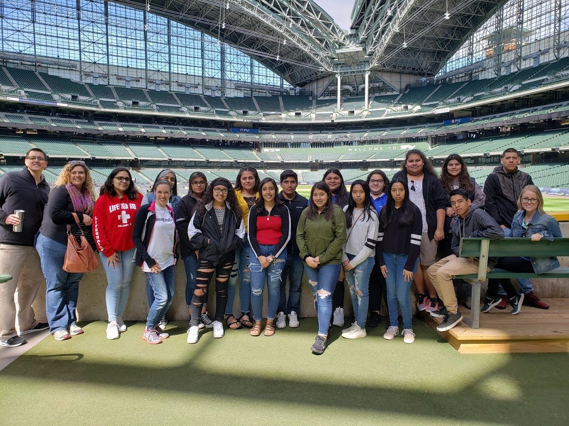 Group of teenagers and adults standing inside a large sports stadium with empty seats and a glass ceiling, posing for a photo.
