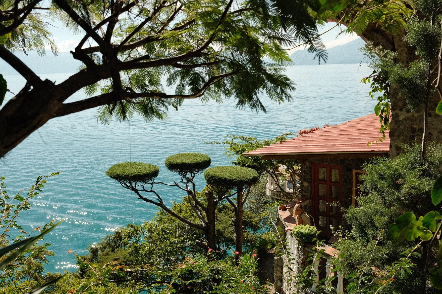 A woman standing on a garden balcony overlooking a lake, surrounded by lush trees and plants, with a stone house nearby.