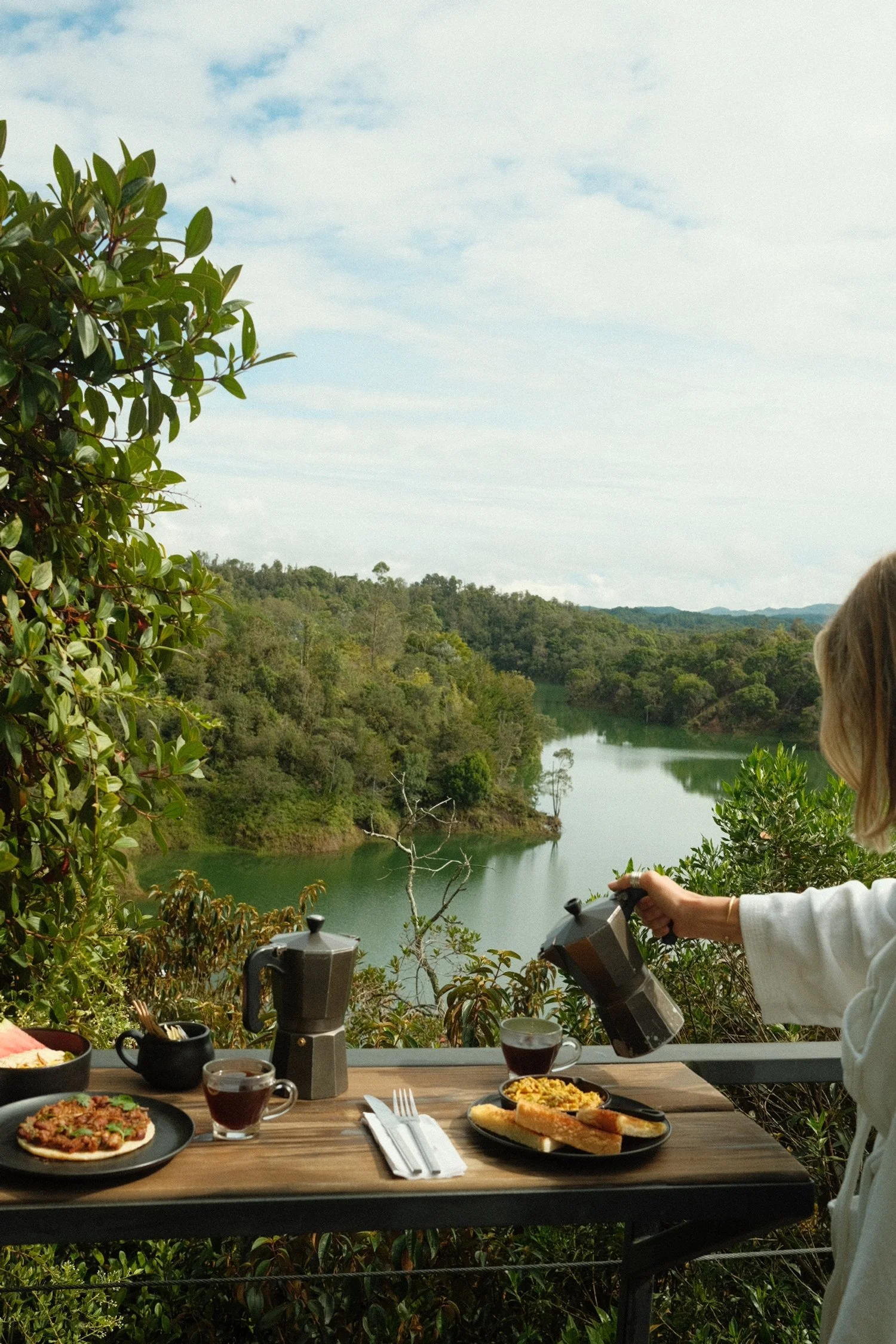 Person pouring coffee on outdoor breakfast table overlooking a river and forested hills under a partly cloudy sky.