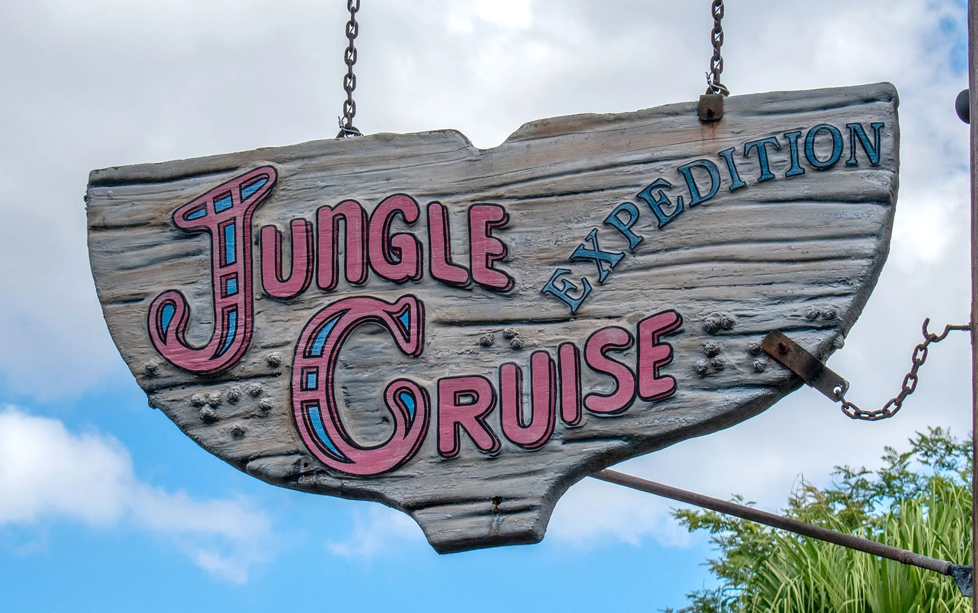 A wooden sign with painted pink and blue letters reads 'Jungle Cruise Expedition'. The sign is weathered and hangs from chains, with a partly cloudy sky and trees in the background.