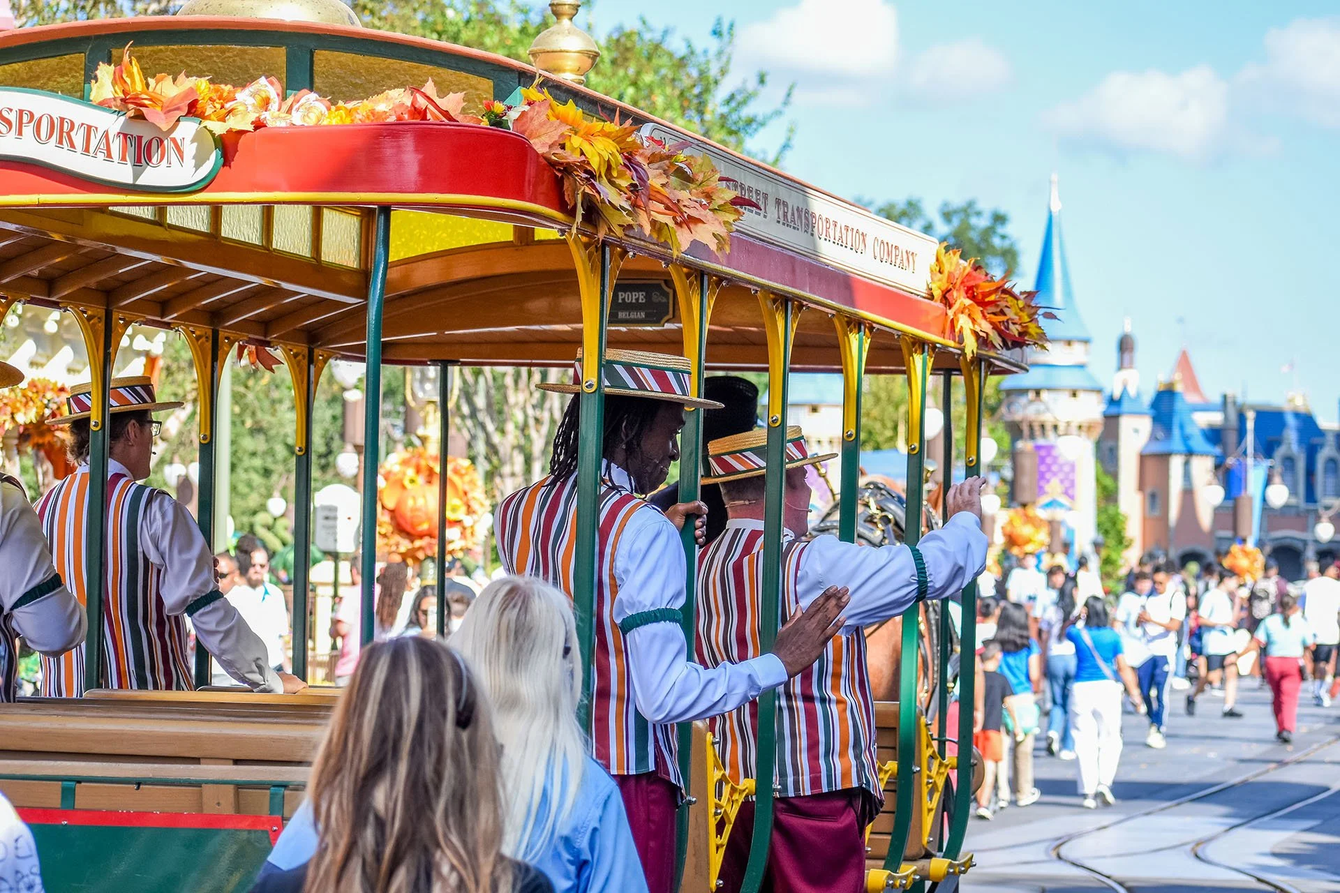 Disney parade featuring a colorful vintage trolley with people waving from it, in front of a whimsical castle and a crowd of guests at Disneyland during daytime.