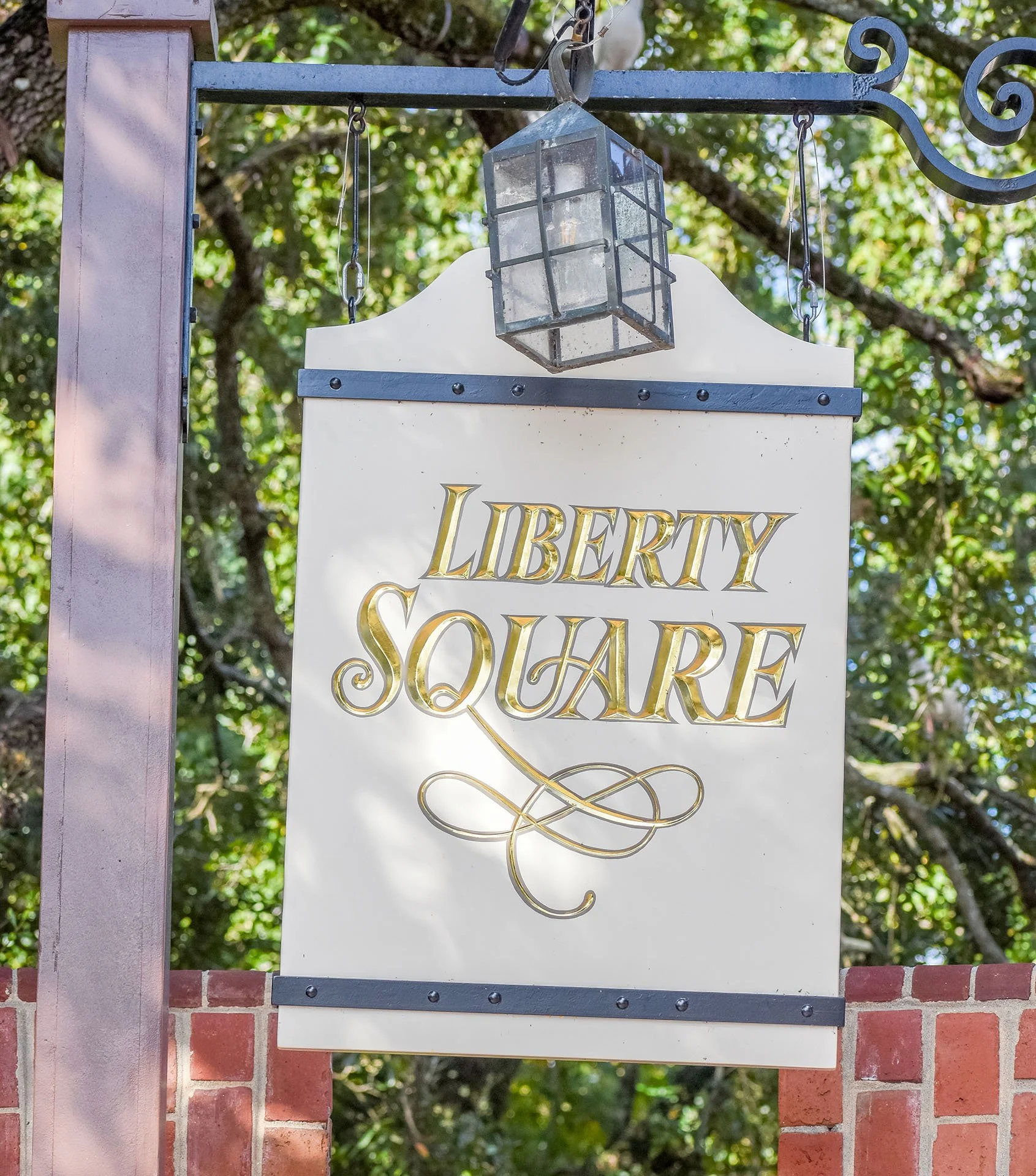 Sign for Liberty Square hanging on a metal frame with a brick wall and green tree in the background.