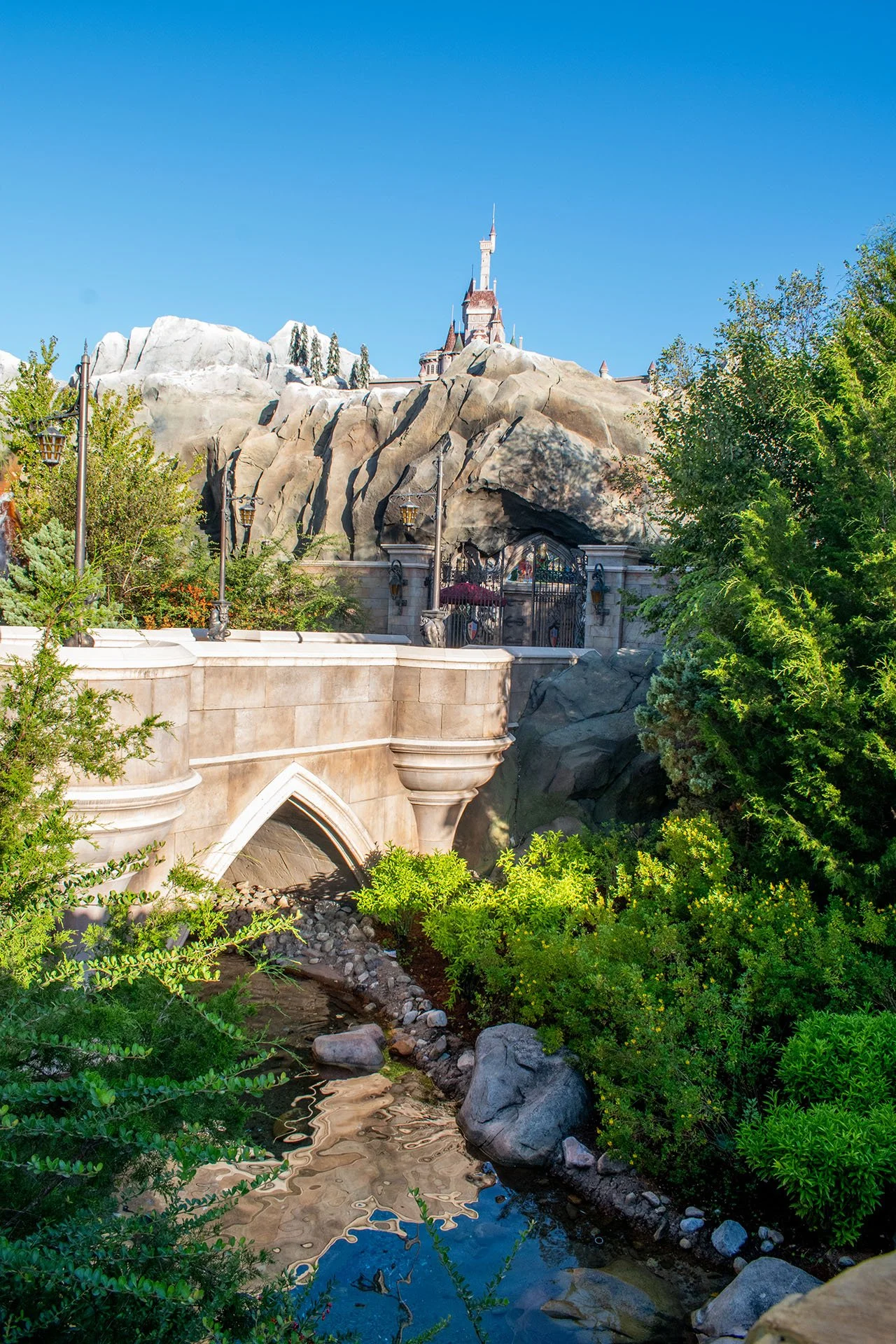 Disney's Sleeping Beauty Castle on a mountain with green trees and a small creek in the foreground, clear blue sky.