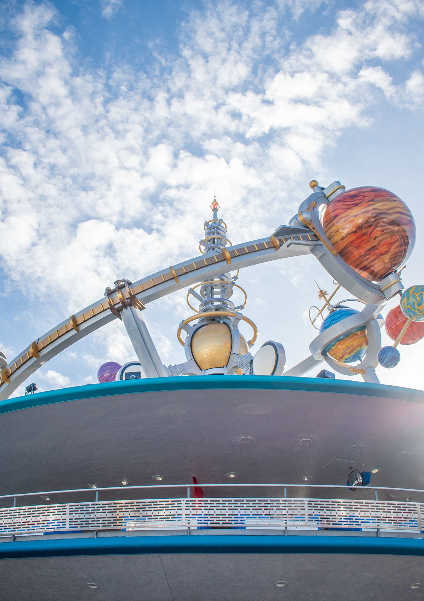 Looking up at a spaceship-themed attraction on a cruise ship deck with planets and stars decor, under a partly cloudy sky.