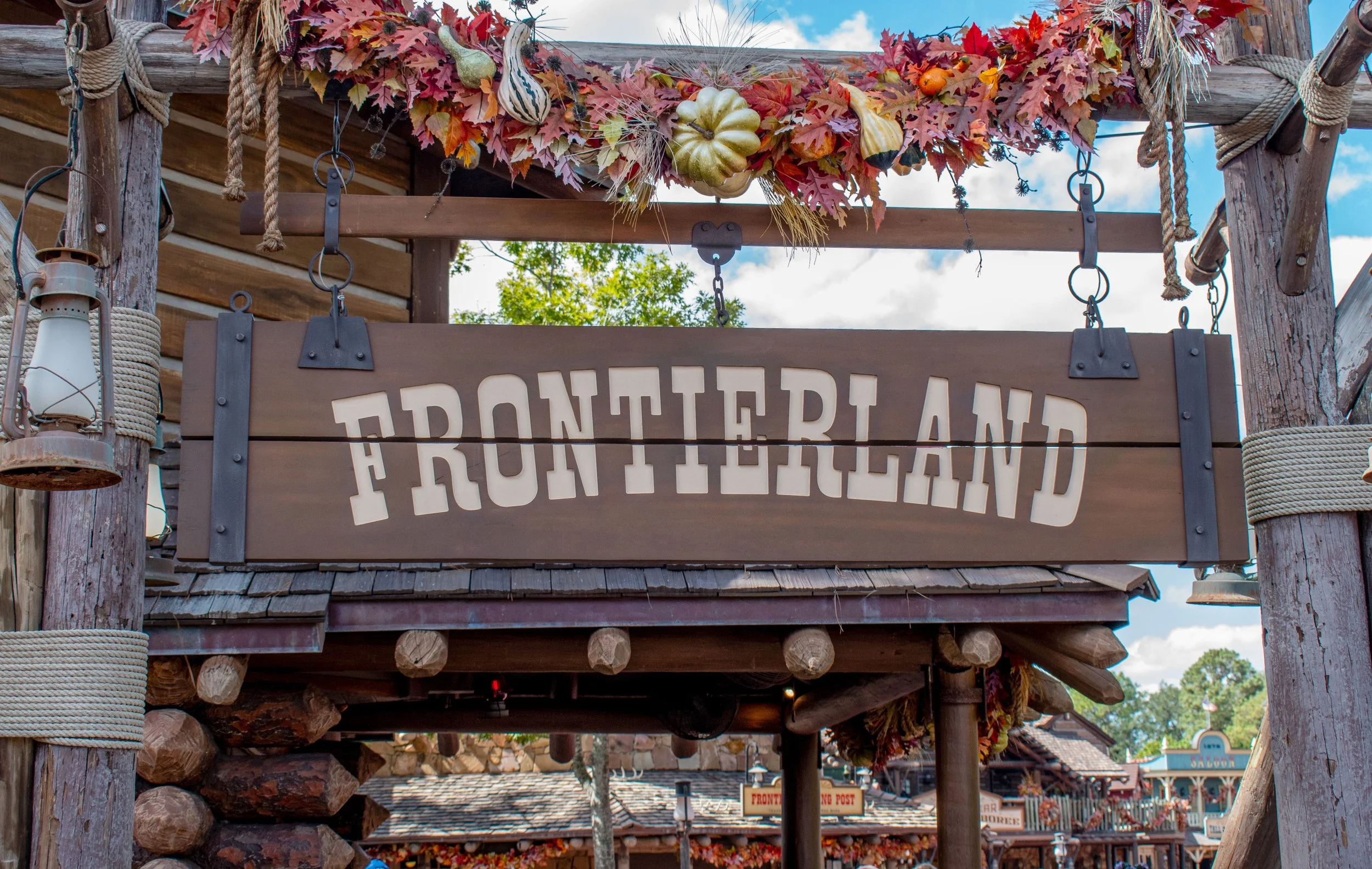 Wooden sign with the word 'Frontierland' hanging above a Western-themed street in a theme park, decorated with fall leaves and pumpkins.