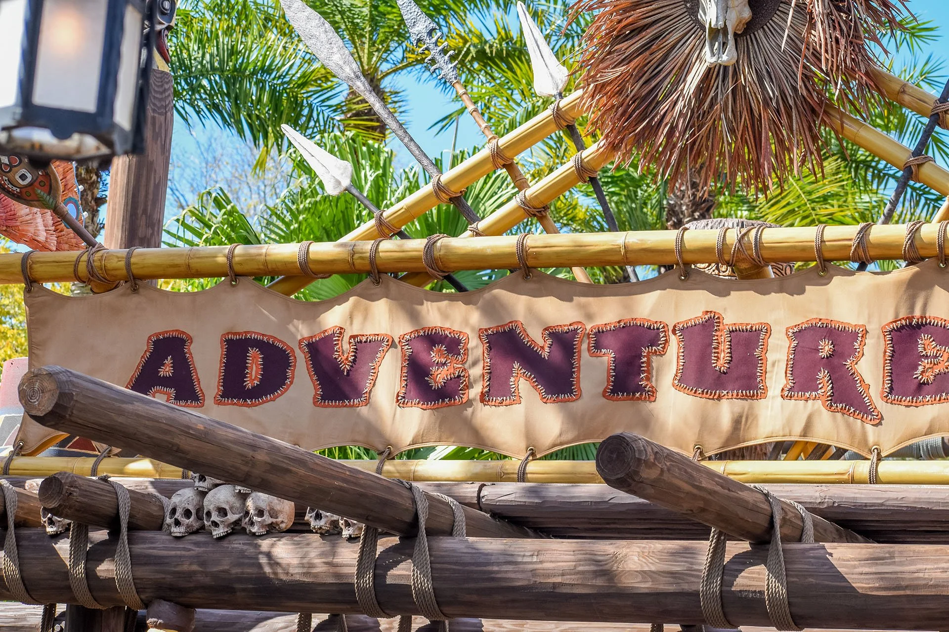 A decorative sign made of fabric and bamboo that reads 'ADVENTURE' with pirate-themed decorations, including skulls and spears, against a background of tropical palm trees and a clear blue sky.