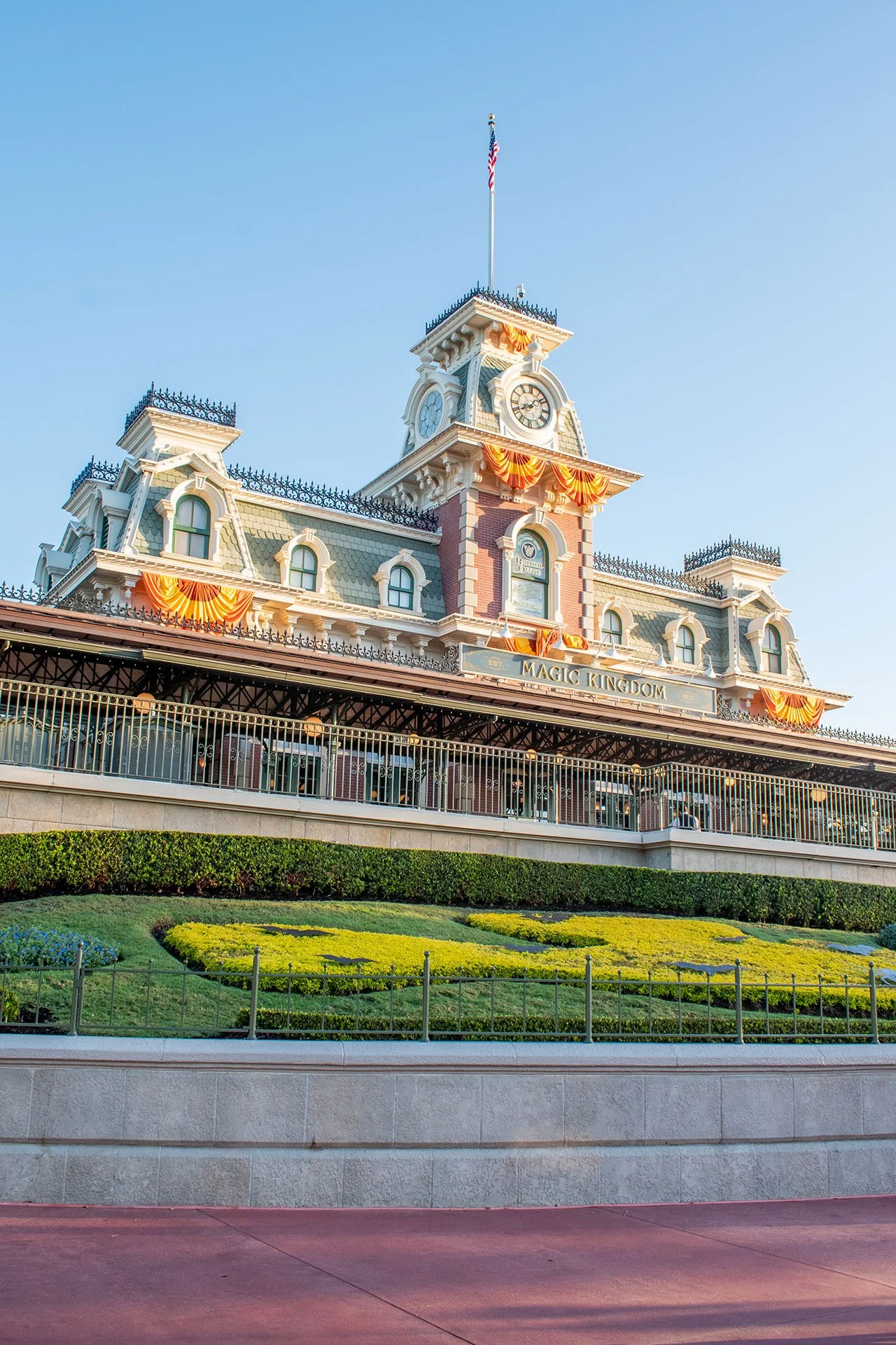 Entrance to Magic Kingdom, a Disney theme park, with a large ornate building featuring a clock tower and a flagpole, decorated with orange and yellow banners, under a clear blue sky.