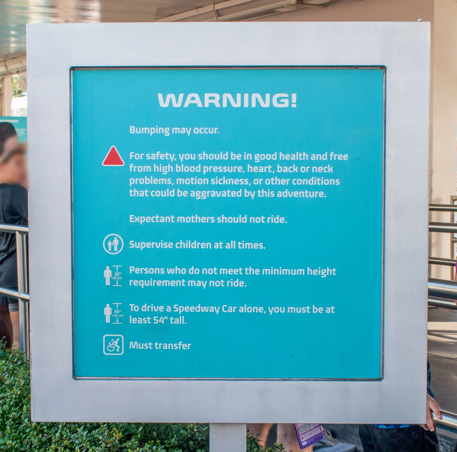 A safety warning sign with instructions for a ride, including health requirements, supervision, height restrictions, and transfer needs, on a blue background with safety symbols.