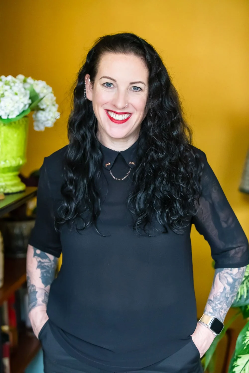 A smiling woman with long curly black hair, wearing a black top with lace sleeves, standing in front of a yellow wall, with a green vase of white flowers on her left.