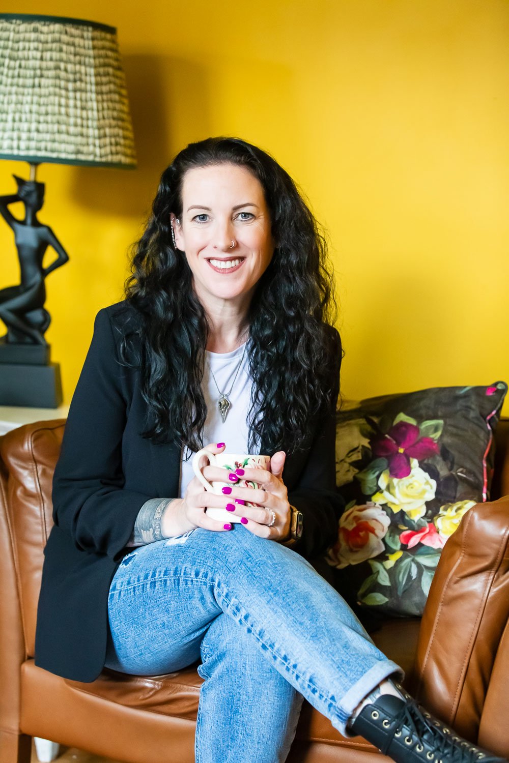 A woman with long, curly black hair, fair skin, and a nose ring, smiling while sitting on a brown leather couch with crossed legs, holding a floral mug, in a room with yellow walls, a decorative lamp, and a floral pillow.