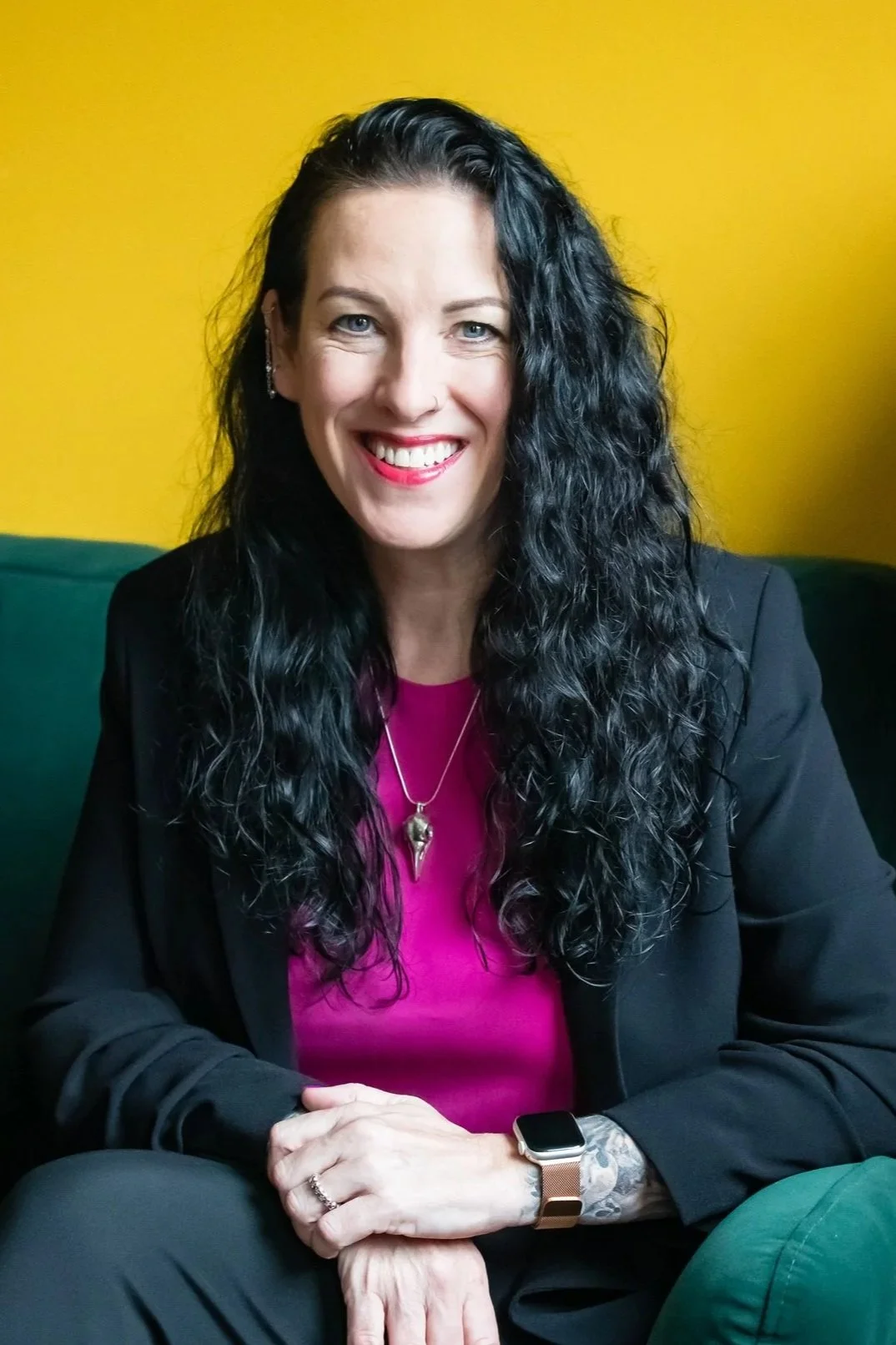 A woman with long, curly black hair, wearing a black blazer over a pink top, smiling while seated on a dark green couch against a yellow wall.