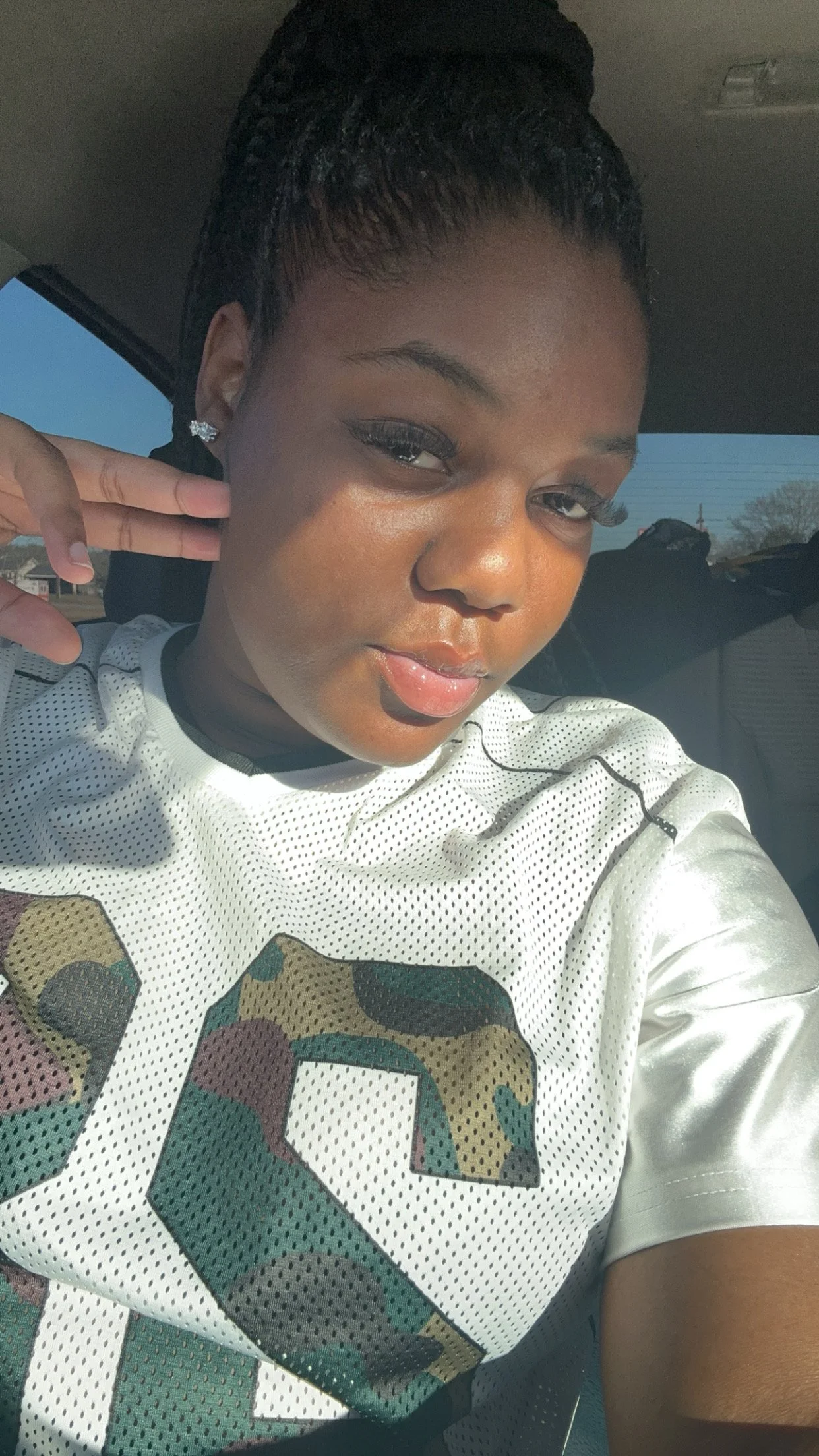 Close-up of a young woman with dark hair styled in a high bun, wearing earrings, makeup, and a white sports jersey with camo pattern on the front, taking a selfie inside a vehicle with bright sunlight coming in.
