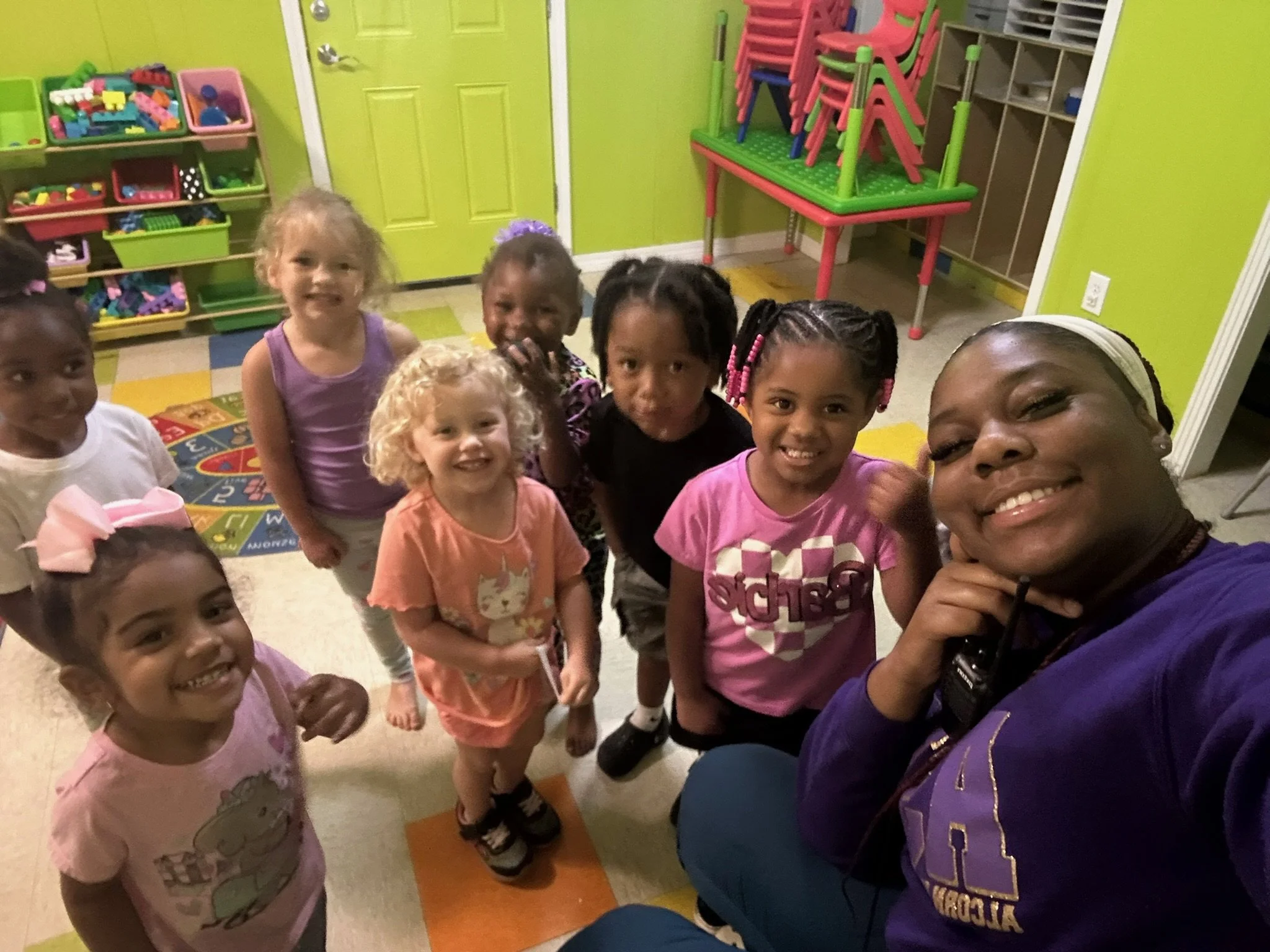 A group of young children and an adult woman smiling and taking a selfie in a colorful classroom with toys and stacked chairs in the background.