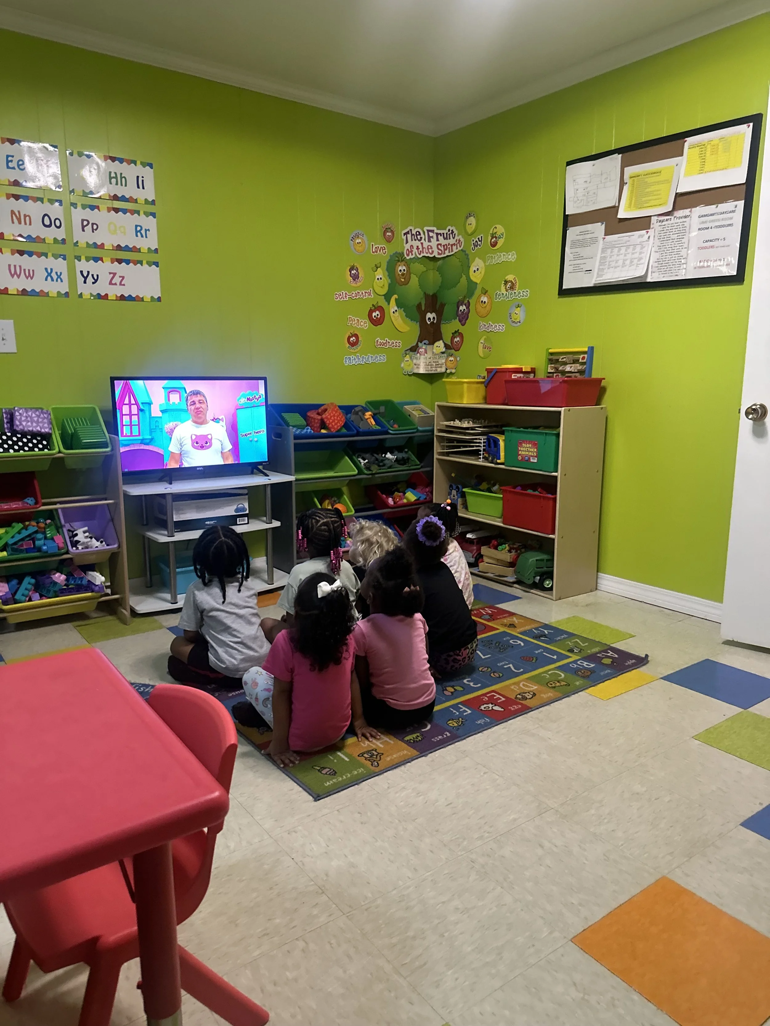Group of children sitting on a colorful alphabet-themed rug, watching a television in a brightly colored classroom with green walls, educational posters, and toy shelves.