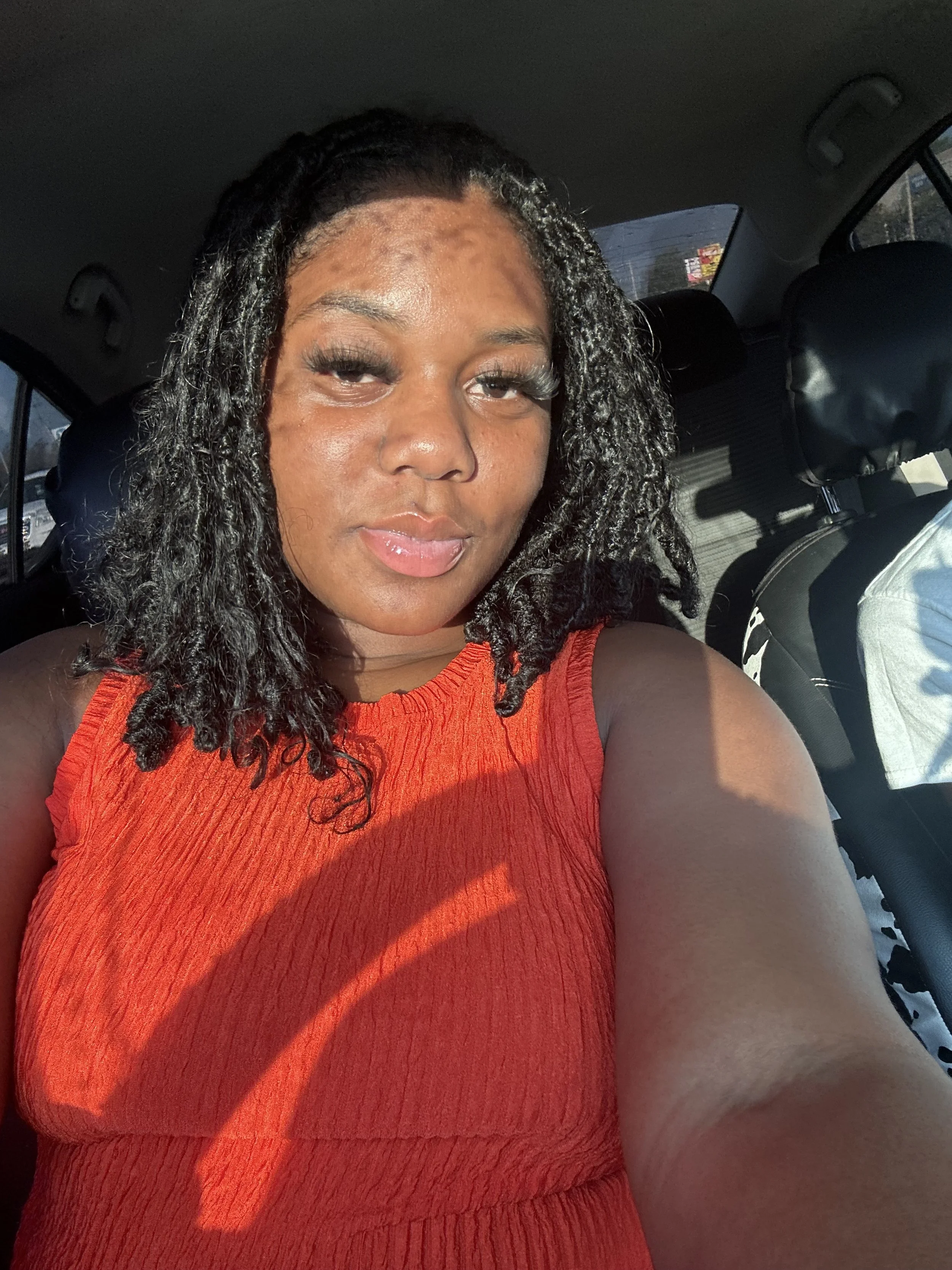 A woman with shoulder-length, curly black hair with white streaks, wearing a sleeveless red top, sitting in a car and taking a selfie.