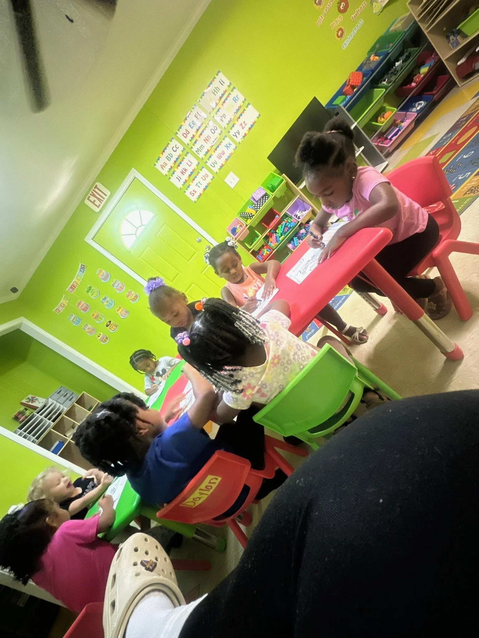 A group of young children sitting at colorful tables in a bright, green-walled classroom, engaged in drawing and coloring activities.