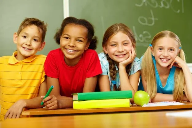 Four smiling school children seated at a desk in front of a chalkboard, with books and an apple in front of them.
