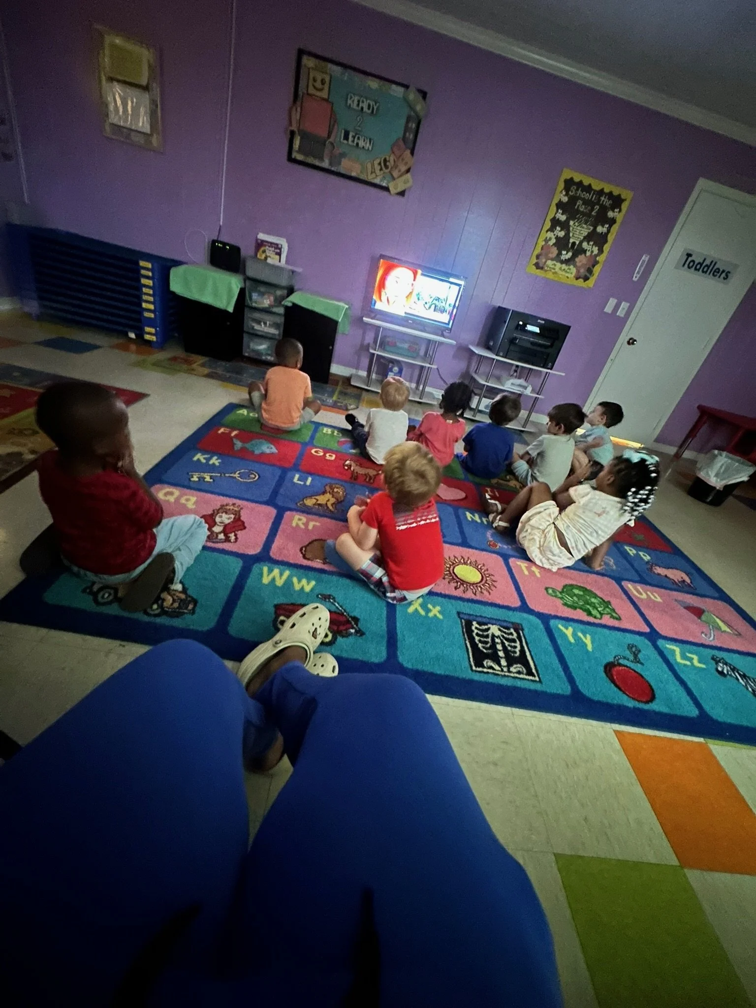 Children sitting on a colorful alphabet rug in a classroom, watching a TV screen.
