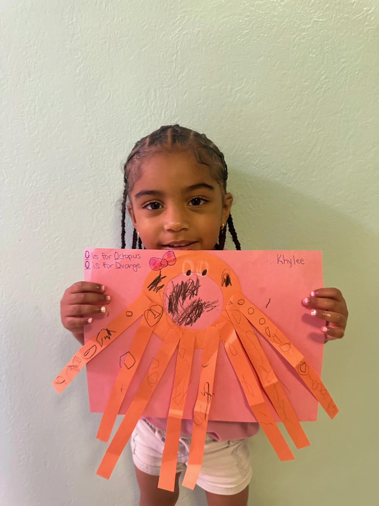 Young girl holding up a pink poster with a colorful octopus drawing made from cut-out orange paper strips, labeled with letters for octopus and orange, on a light green wall background.