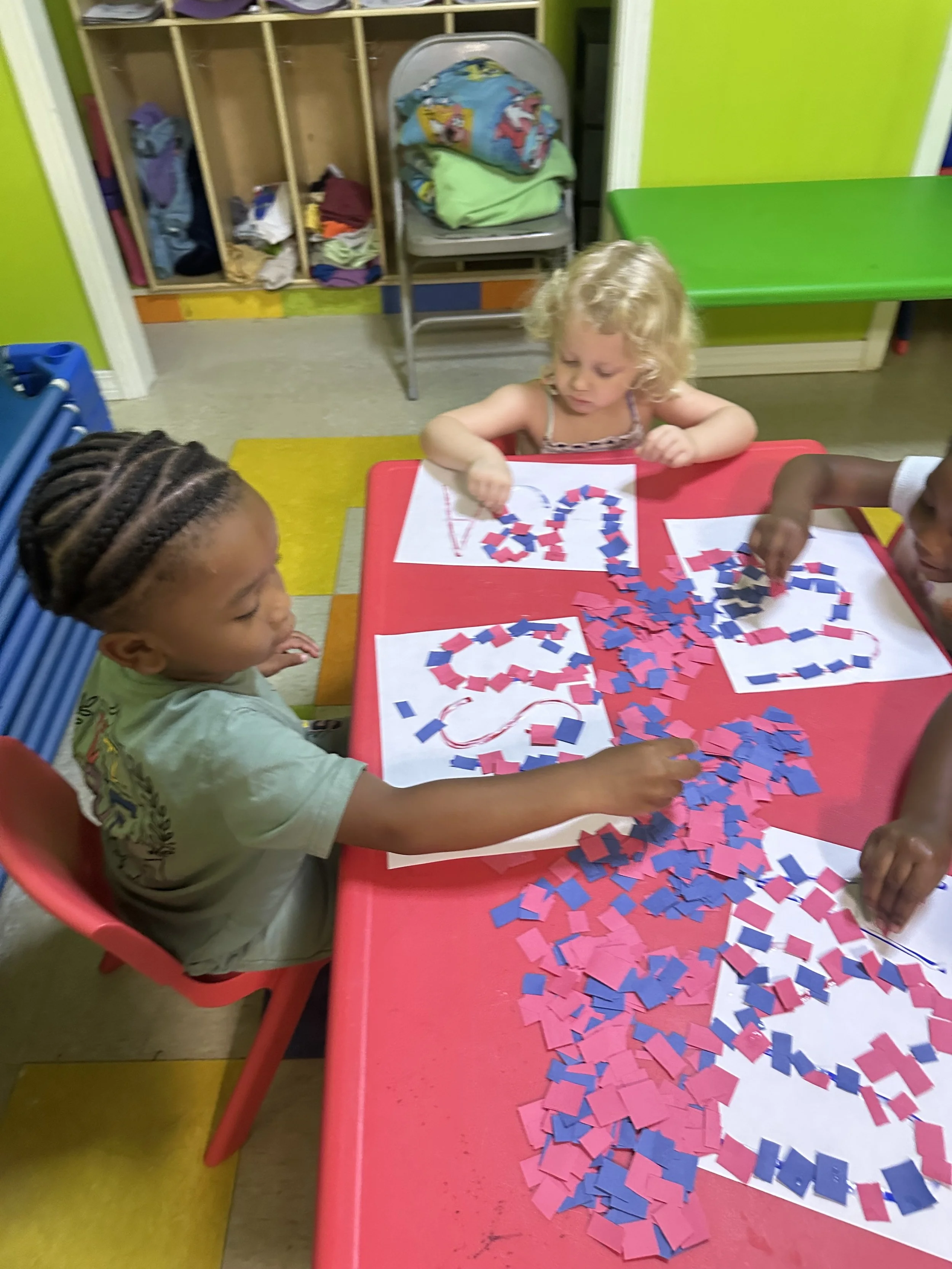 Children sitting at a red table, creating artwork with red, blue, and pink paper shapes on white paper, in a colorful classroom.