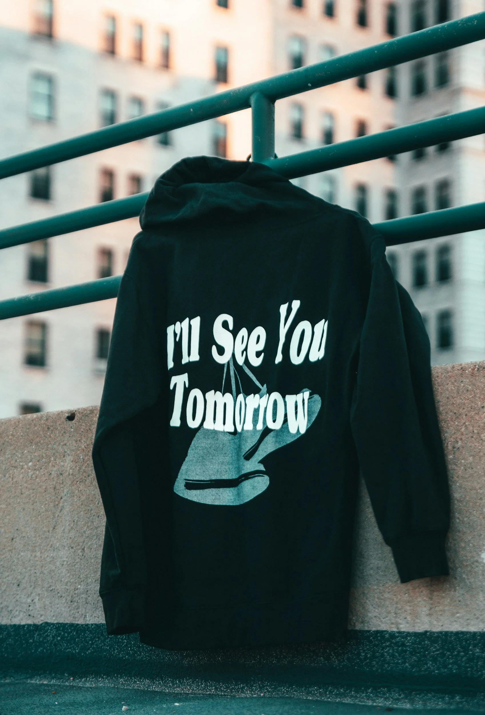 Black hoodie hanging on a green metal railing in an urban setting with tall buildings in the background. The hoodie features white text that says, "'Til See You Tomorrow" with a graphic of a hand holding a cigarette.
