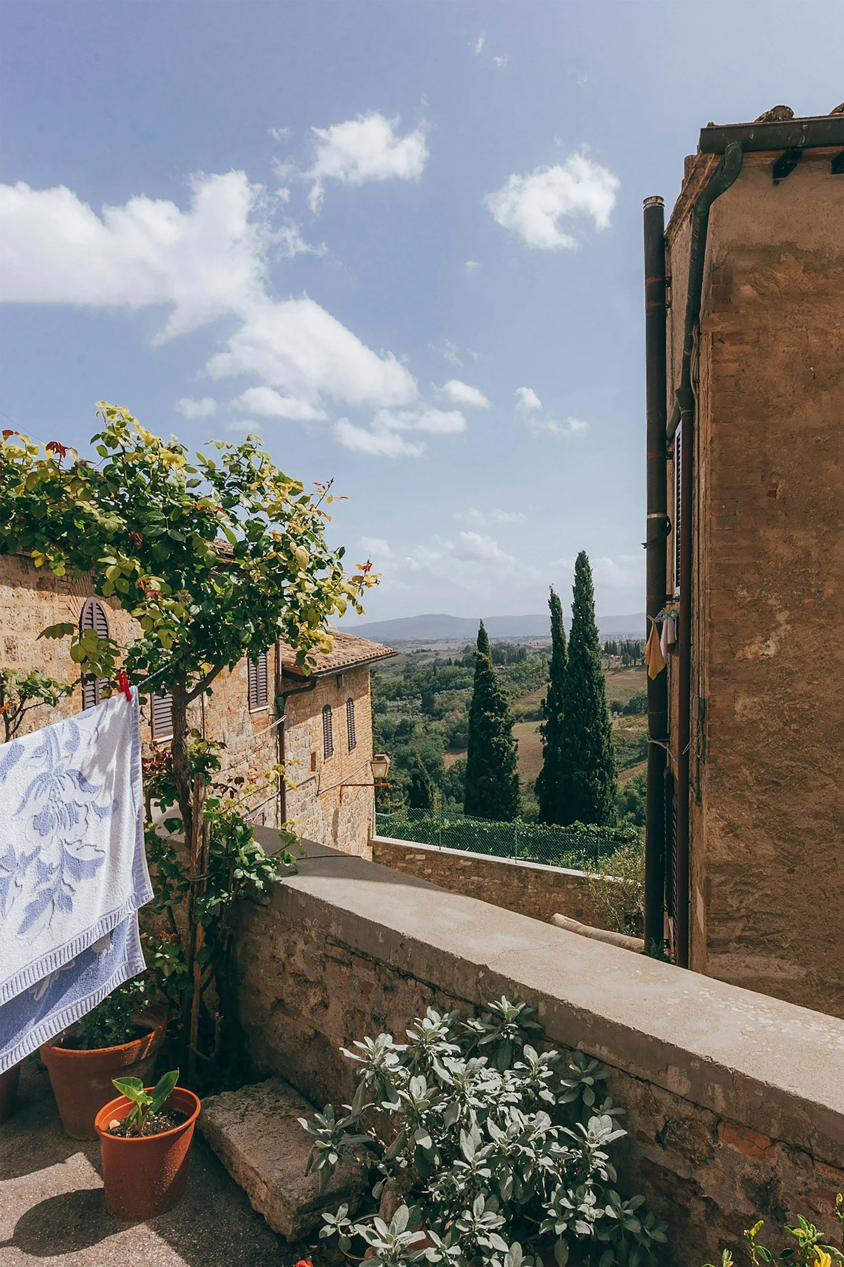 A rustic balcony with potted plants, a towel hanging on a clothesline, overlooking rolling hills and tall cypress trees under a partly cloudy sky.