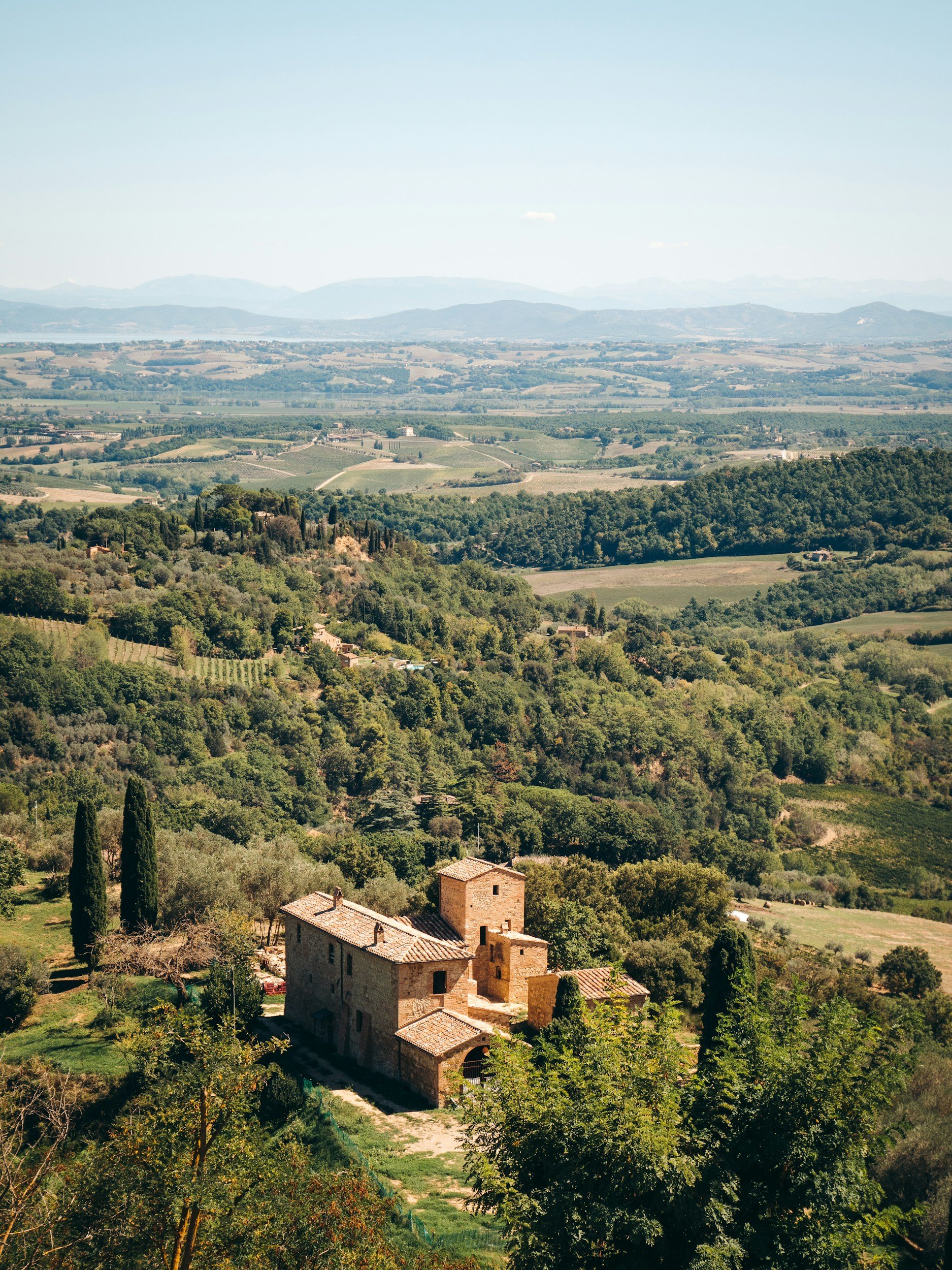 Scenic view of a rustic stone house in the Tuscan countryside surrounded by greenery, rolling hills, and distant mountains under a clear blue sky.