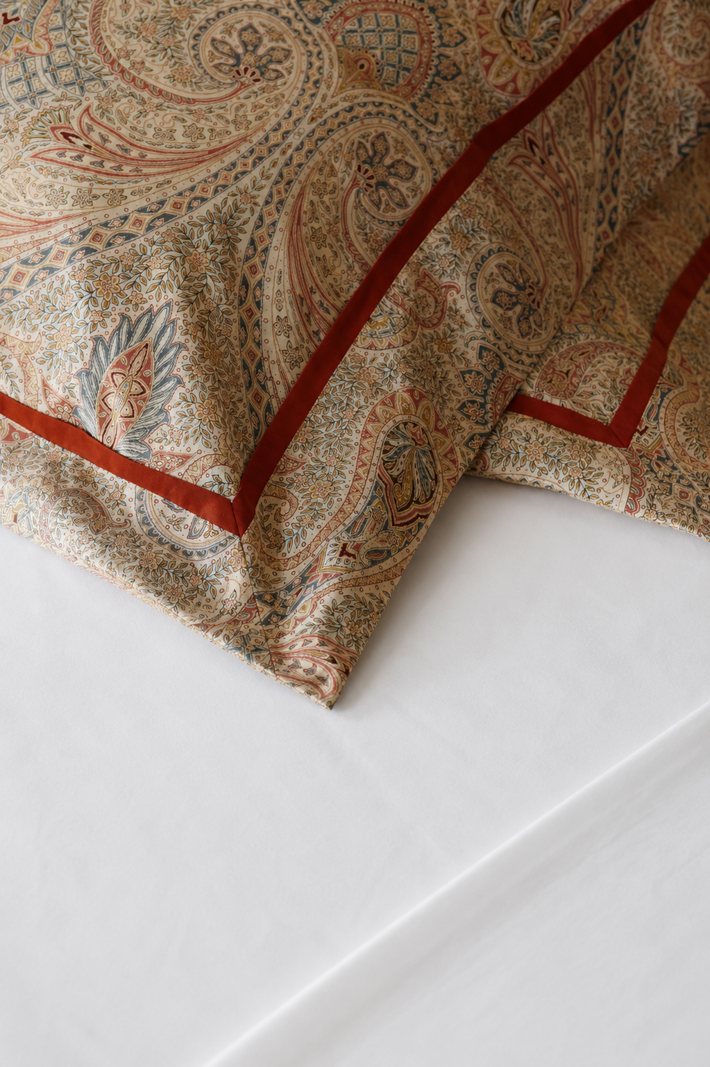Close-up of a decorative pillow with intricate paisley and floral patterns in beige, red, blue, and gold, resting on a white bed sheet.