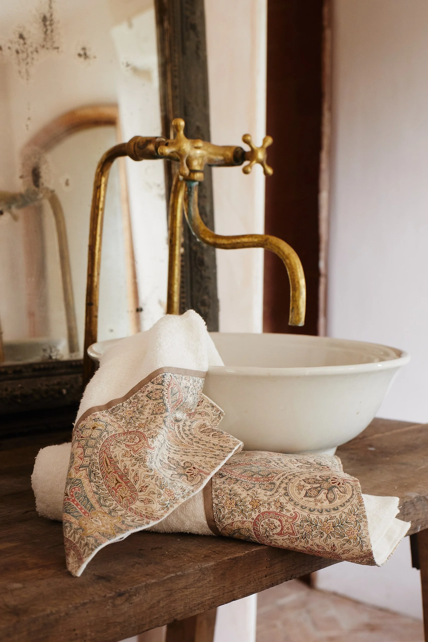 A vintage brass faucet above a white ceramic washbasin on a rustic wooden table, with a rolled white towel and a decorative fabric napkin placed nearby.