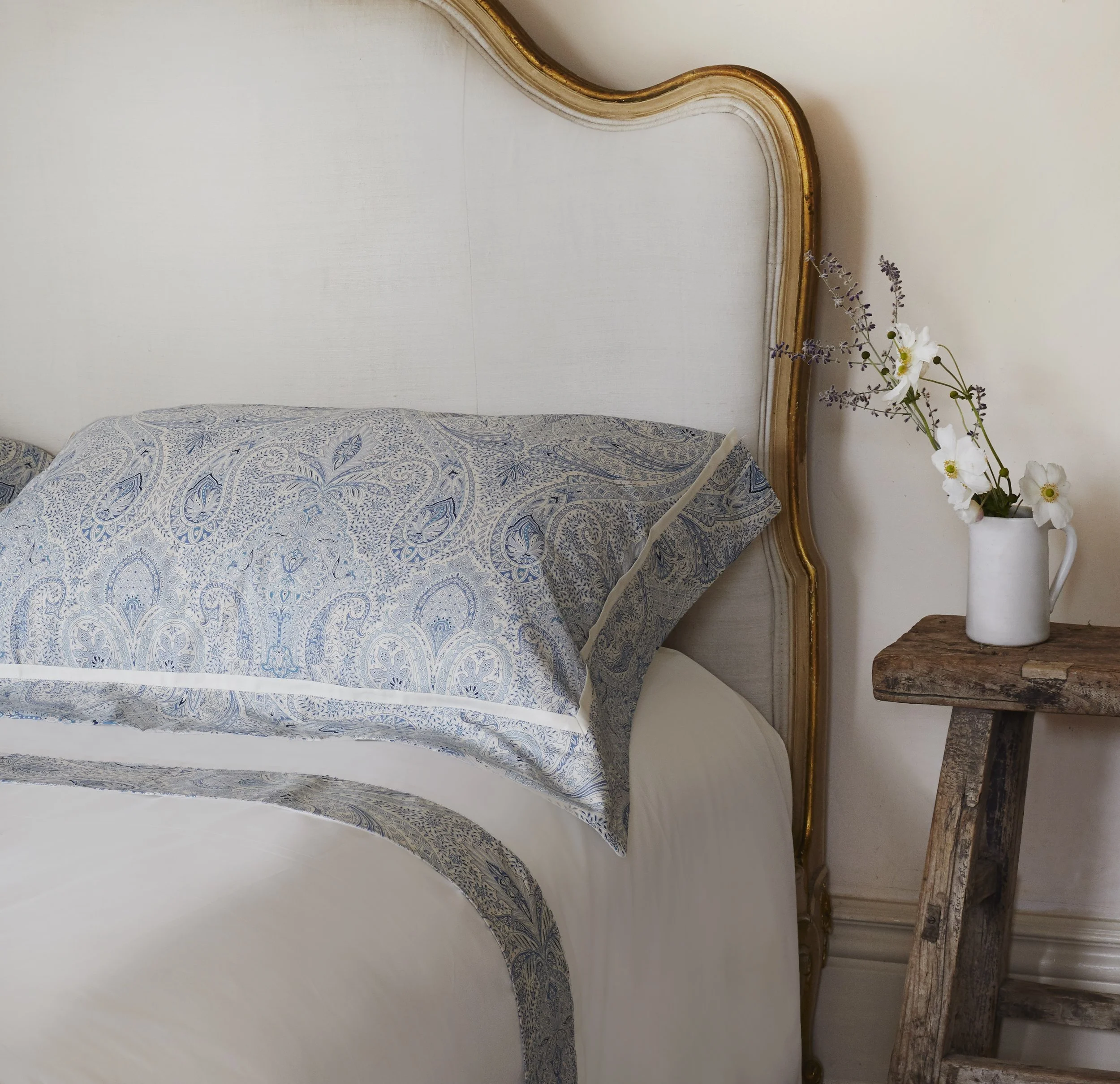 A bedroom with a gold-trimmed headboard, a pillow with a blue and white paisley pattern, and a wooden nightstand holding a white vase with white and purple flowers.