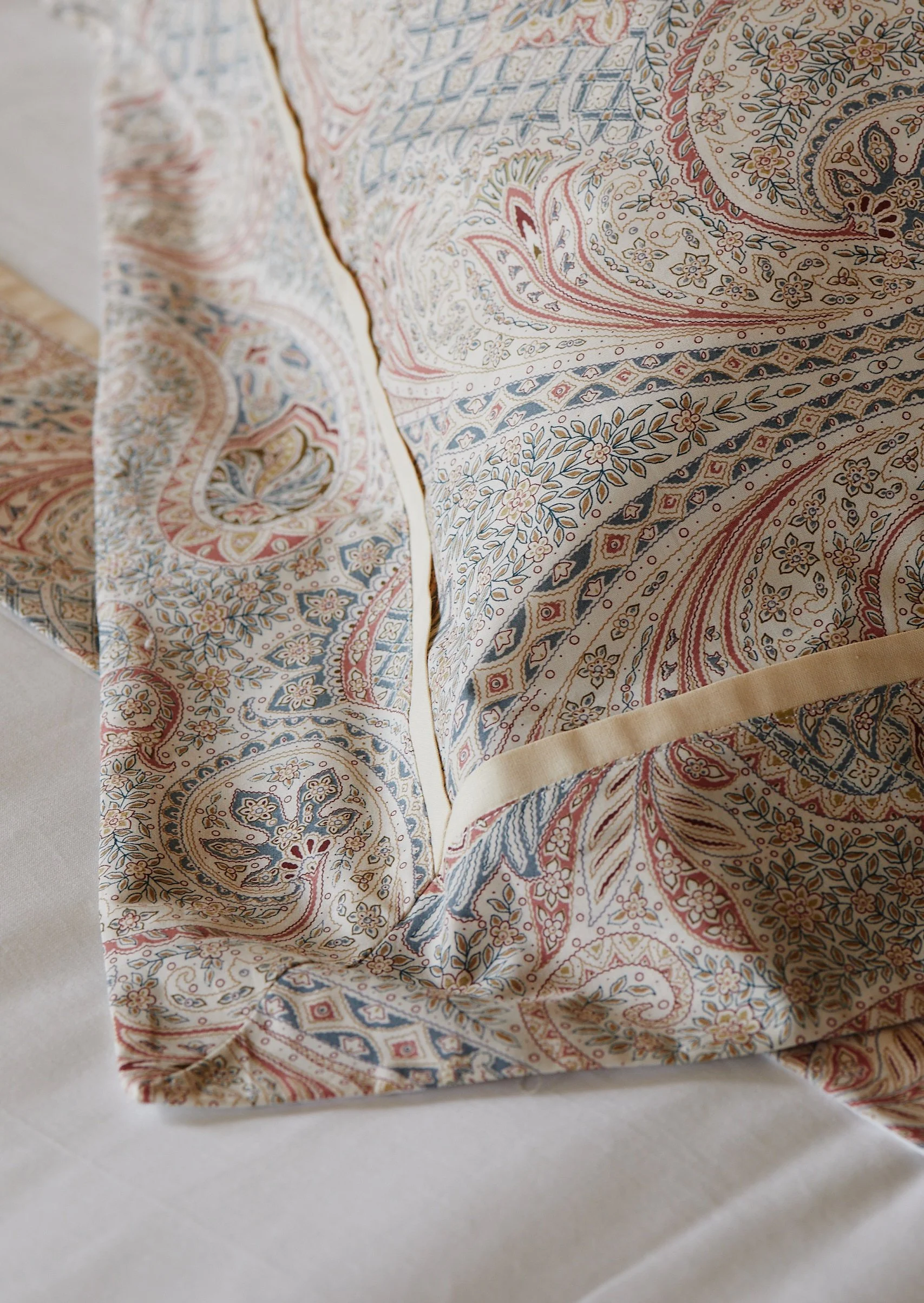 Close-up of a decorative fabric with intricate paisley and floral patterns in beige, red, blue, and gold, draped over a white surface.