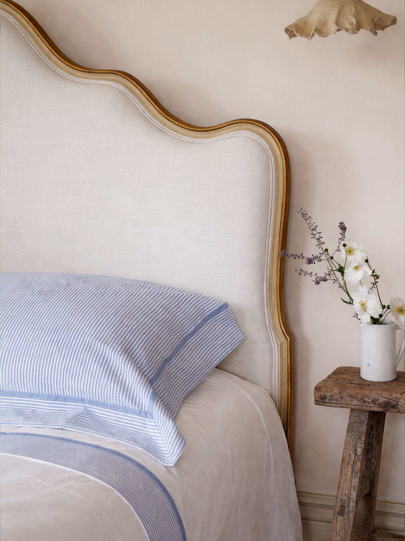 A bedroom corner with a white upholstered headboard with wooden trim, a striped blue and white pillow, and a rustic wooden nightstand with a white vase holding white and purple flowers.