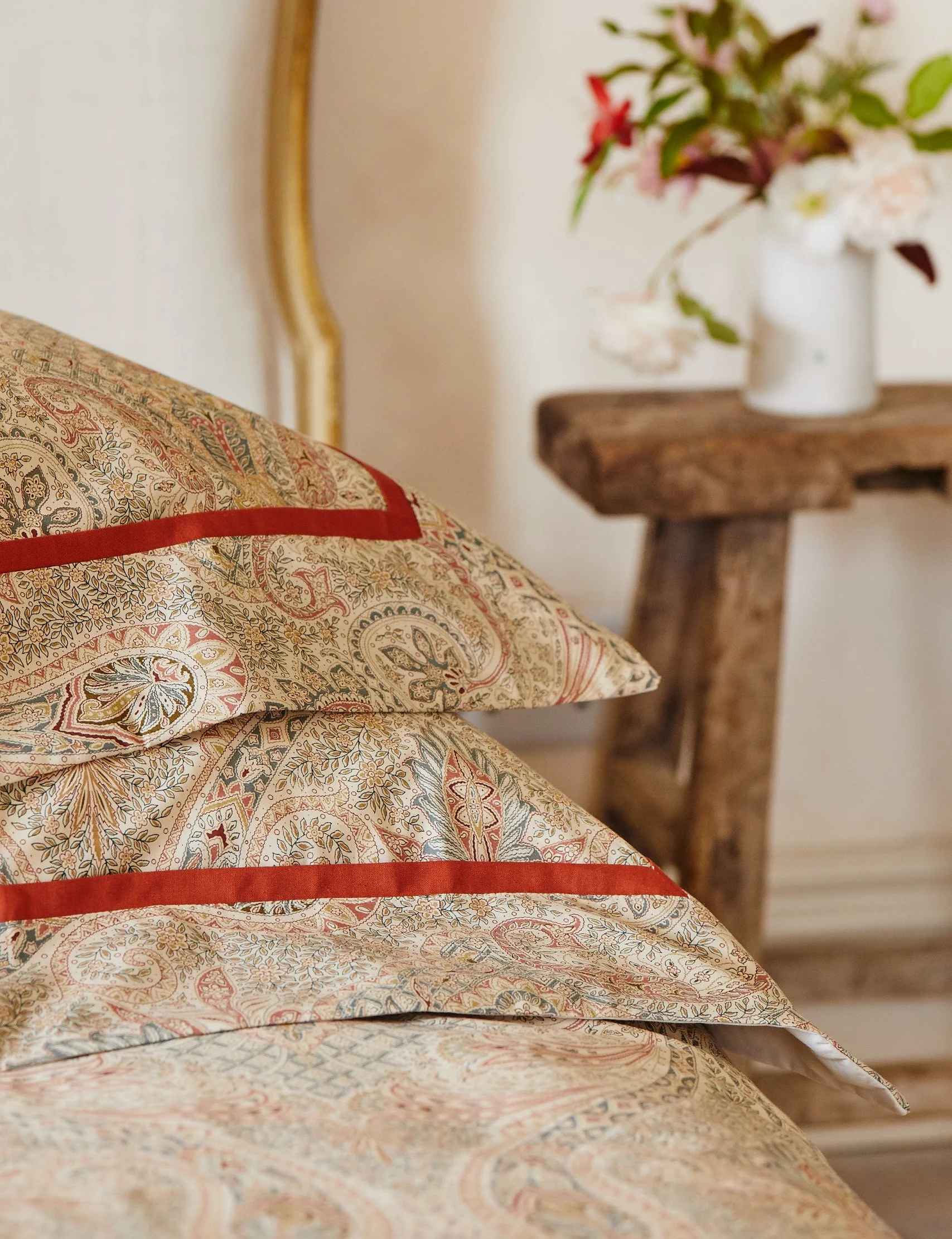 Close-up of decorative pillows with intricate paisley and floral patterns on a bed, with a rustic wooden bedside table holding a vase of flowers in the background.