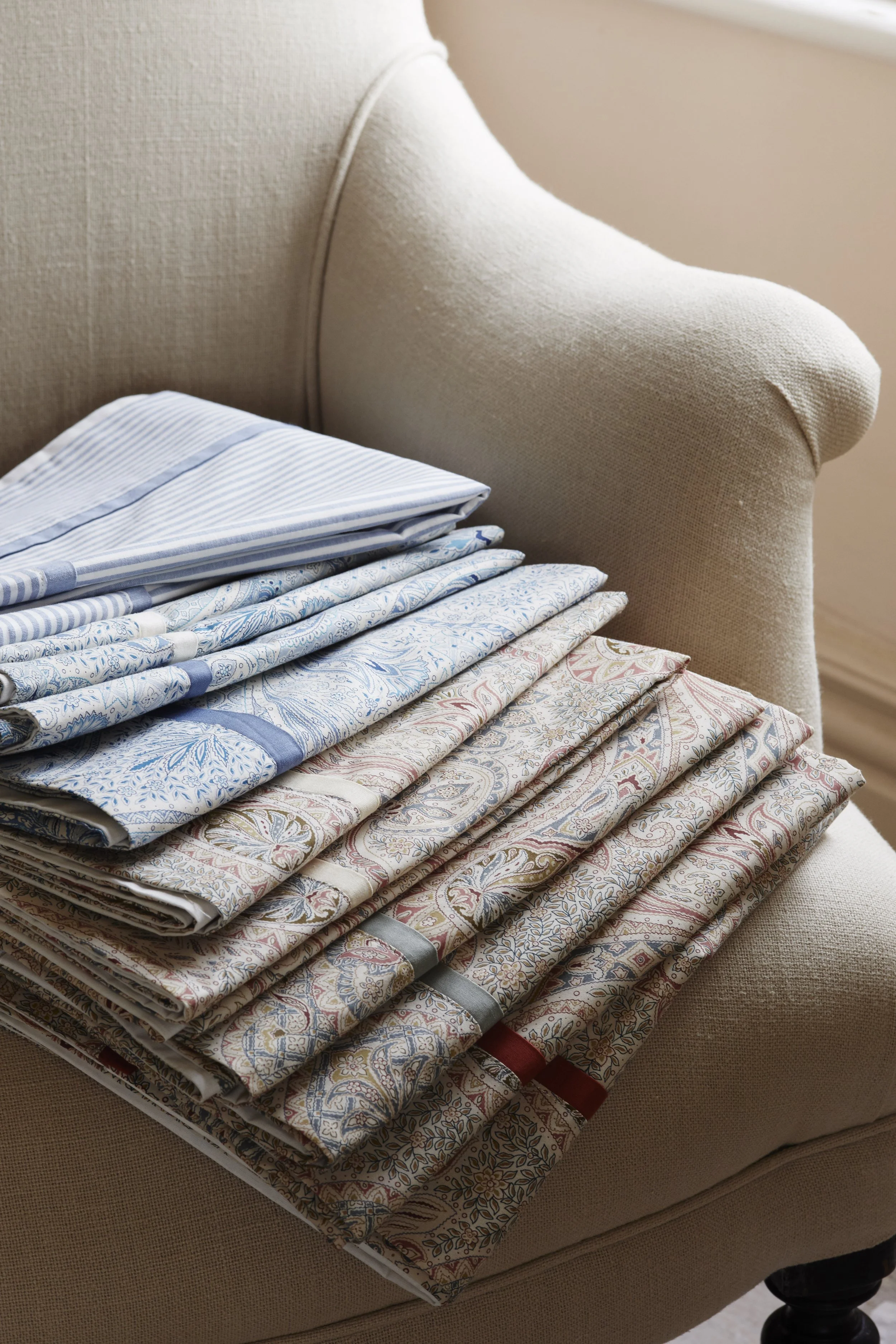 Stacked bed linens with various patterns and colors, placed on a beige upholstered armchair.