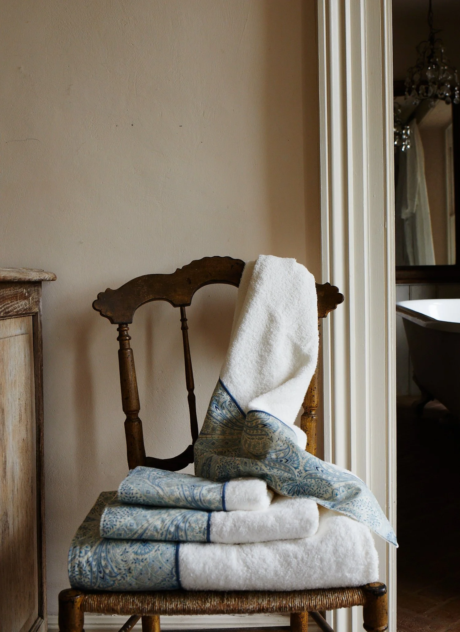 A wooden chair with a white towel and folded blue and white patterned towels on it, situated next to a beige wall and a doorway leading to a bathroom with a clawfoot bathtub.