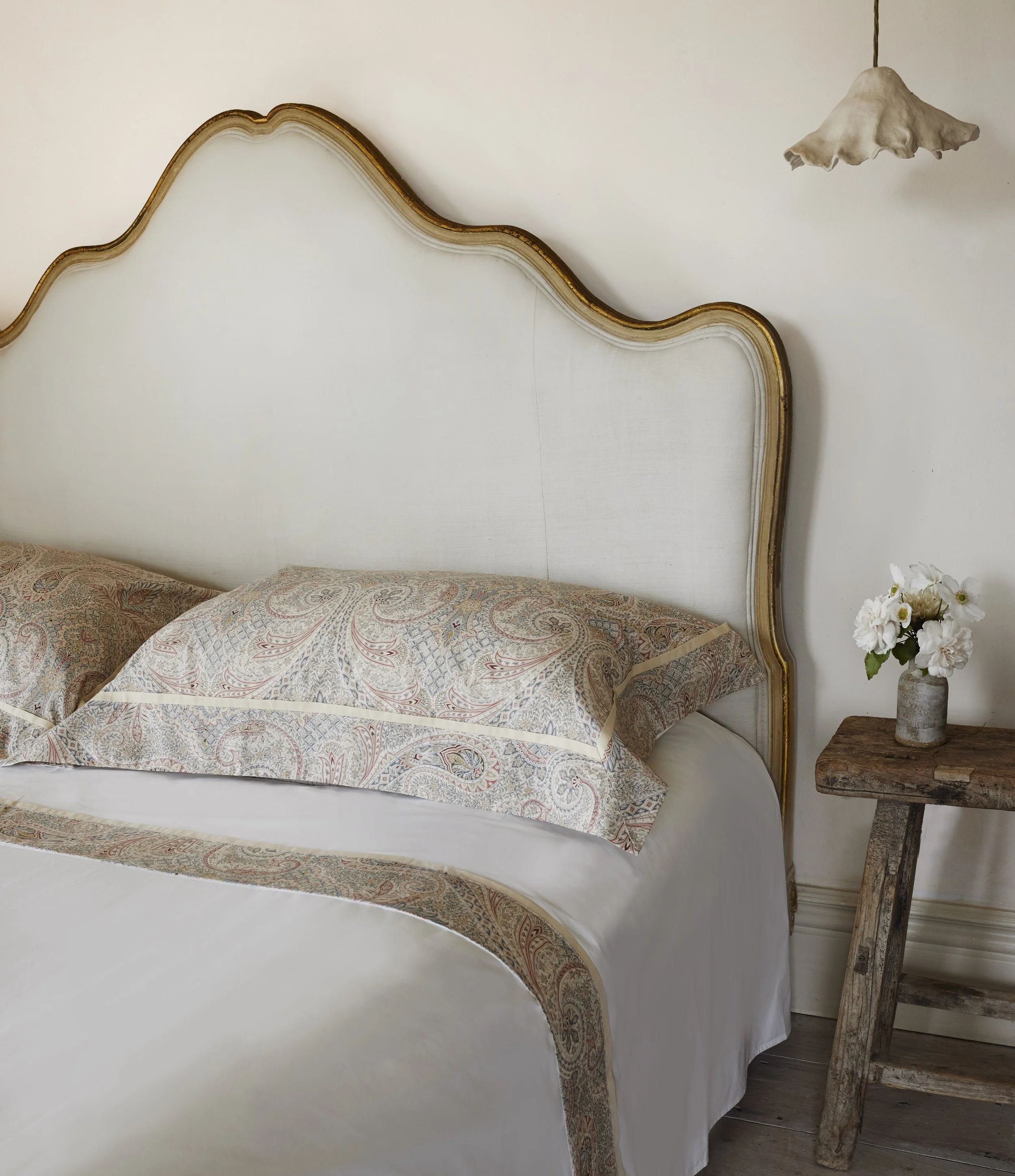 A bedroom with a white bed and a vintage headboard with gold trim, a wooden side table with a vase of white flowers, and a ceiling lamp with a wavy, beige lampshade.