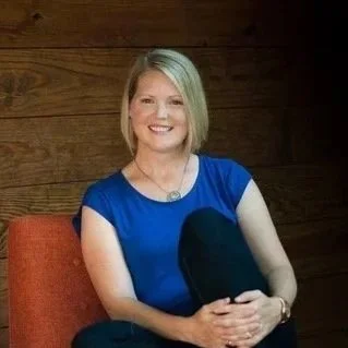 Angela Schaffner sits on red couch, in a blue shirt. She is blonde and smiling/